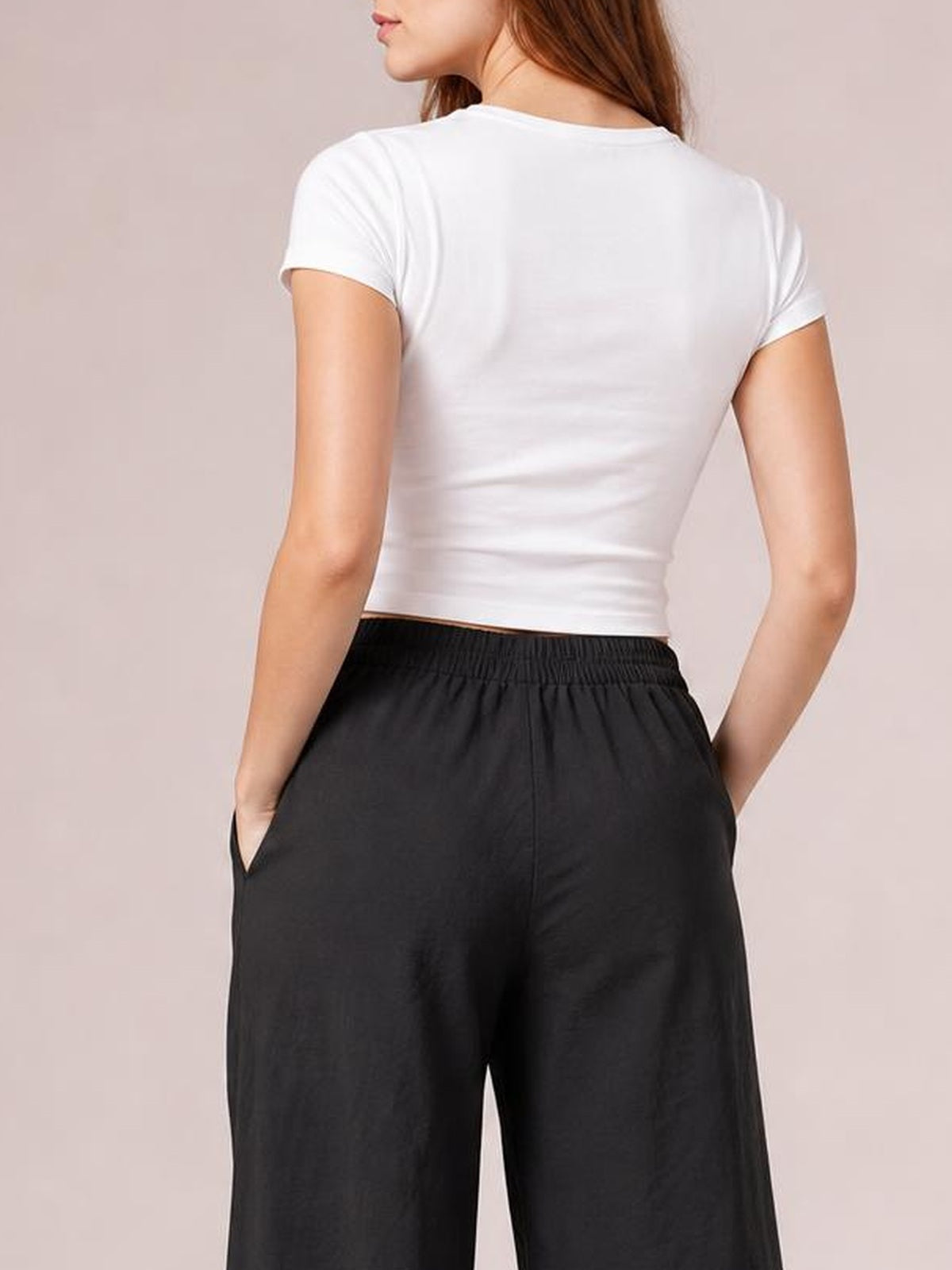 Model seen from behind wearing black wide leg pants with an elastic waistband and side pockets, standing with both hands tucked into the pockets.