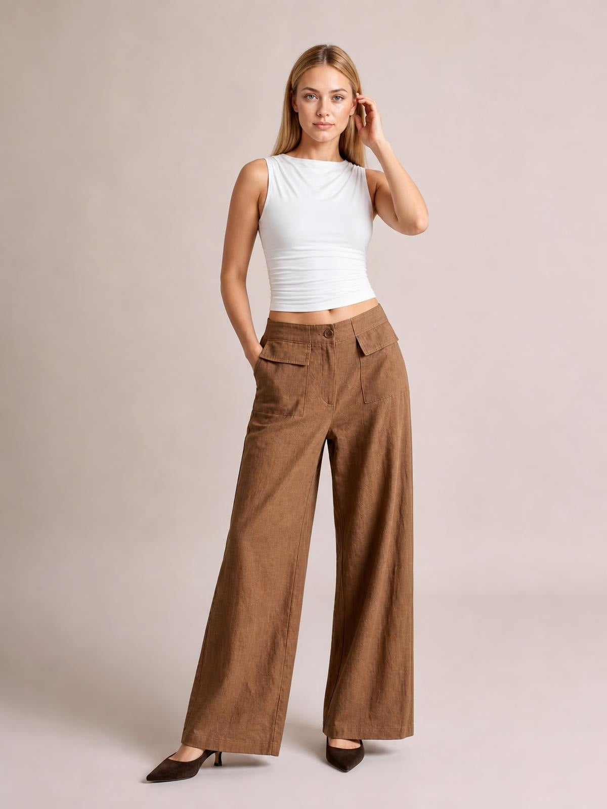 A model poses with one hand in a pocket and the other touching her hair, wearing brown wide leg pants with a waistband, button closure, patch pockets, and relaxed legs.