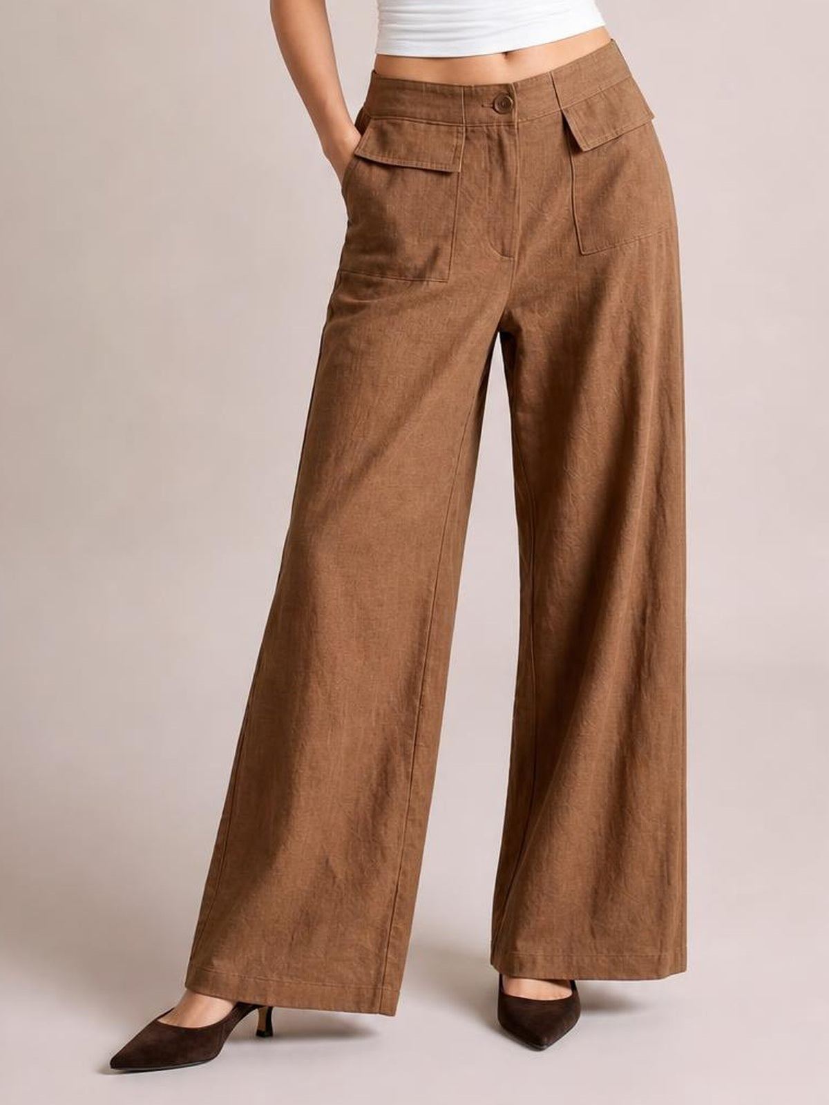 Model poses with one hand in a pocket, wearing brown wide leg pants with a button waistband, front patch pockets, flap pockets, and a relaxed full-length silhouette.