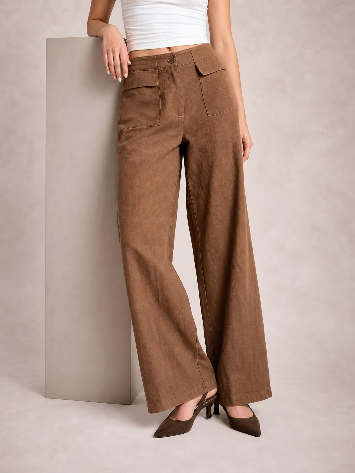 Model leans against a block wearing brown wide leg pants with a buttoned waistband, large front patch pockets, straight seams, and a relaxed full-length silhouette.