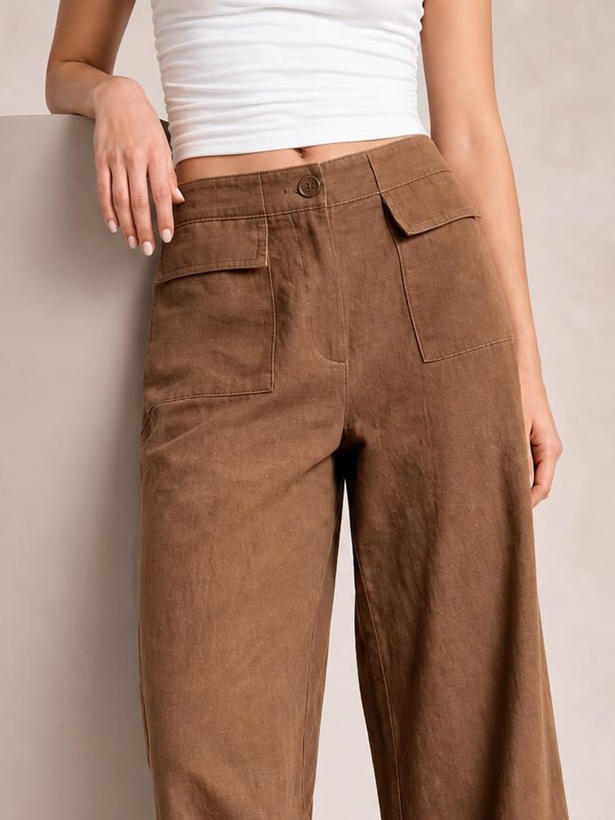 Model in a white top wears brown wide leg pants with a button closure, stitched waistband, and large flap pockets.