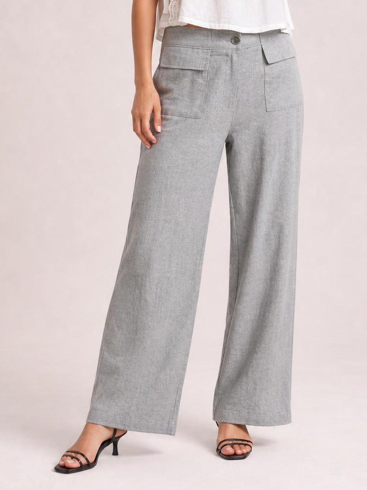 Model wearing light gray wide leg pants with a button closure, belt loops, and large flap pockets, standing in strappy heeled sandals.