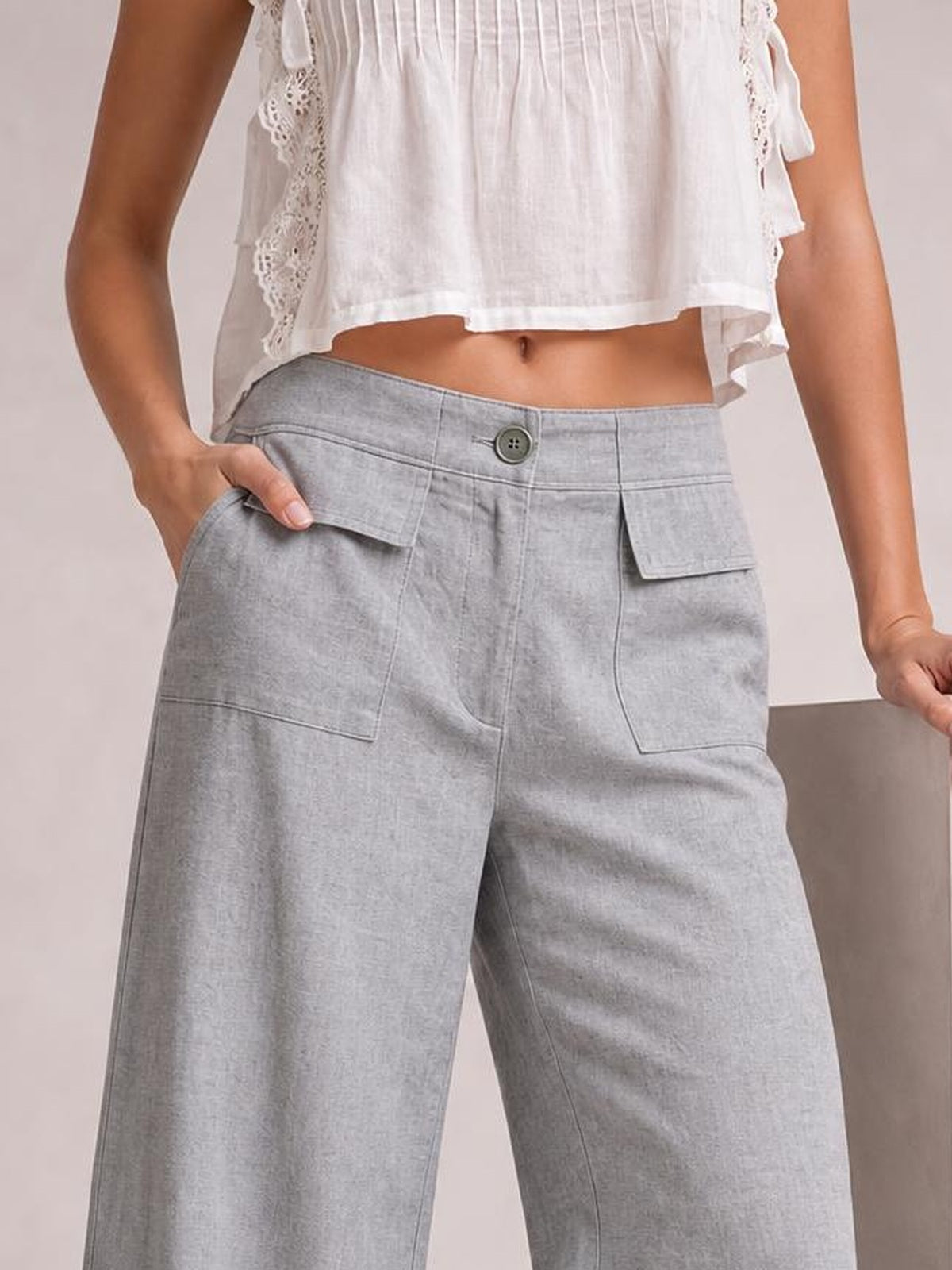 Model wears light gray wide leg pants with a button waistband, front seam details, and flap pockets, posing with one hand in a pocket.