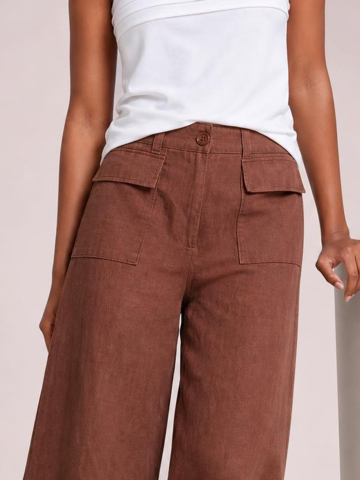 Model wearing brown wide leg pants with a high waistband, button closure, belt loops, and large flap pockets, shown in a close-up standing pose.