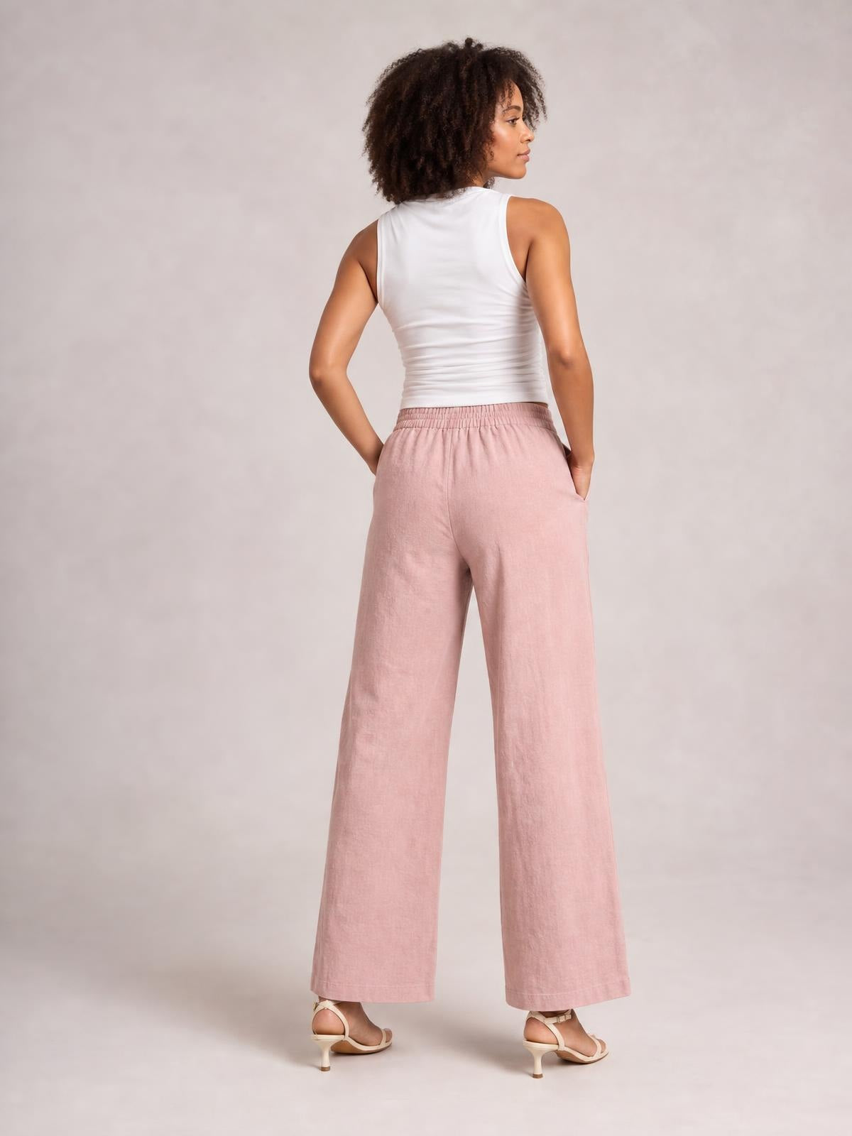 Model shown from behind wears pink wide-leg pants with a gathered elastic waistband and side pockets, standing in a relaxed pose with hands tucked in.