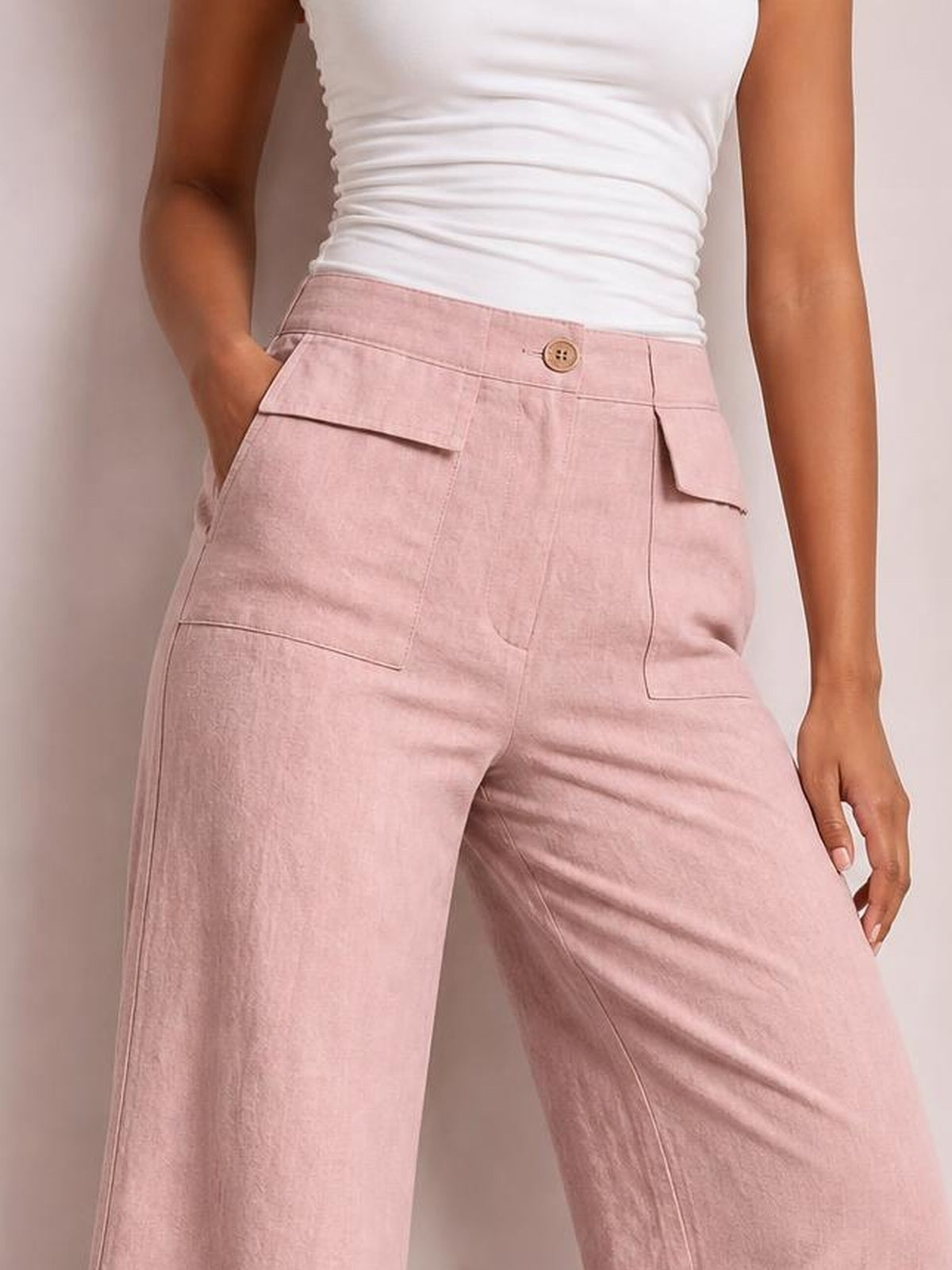Model wearing pink wide leg pants with a high waist, button closure, stitched seams, and flap pockets, posed with one hand in a pocket.