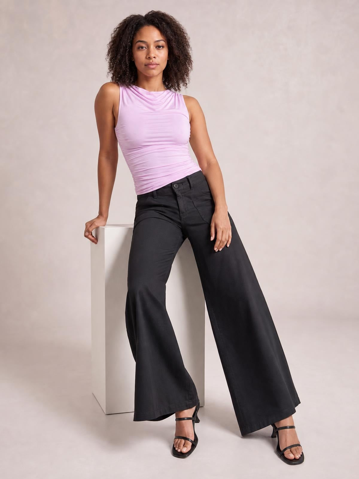 A model leans against a white block wearing black wide leg pants with front pockets, belt loops, button closure, and a flowing full-length silhouette.