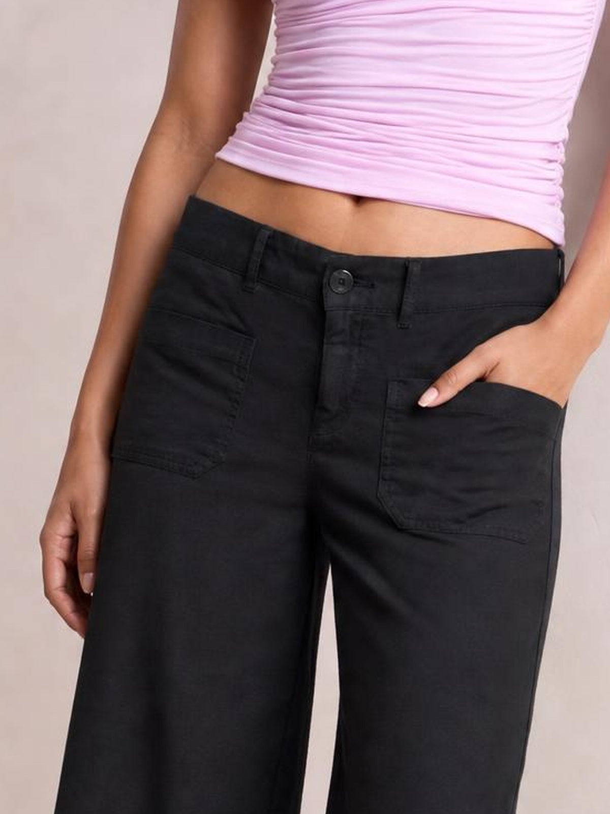 Model wearing black wide-leg pants with front pockets, belt loops, button closure, and stitched pocket detailing, posed with one hand tucked into a pocket.