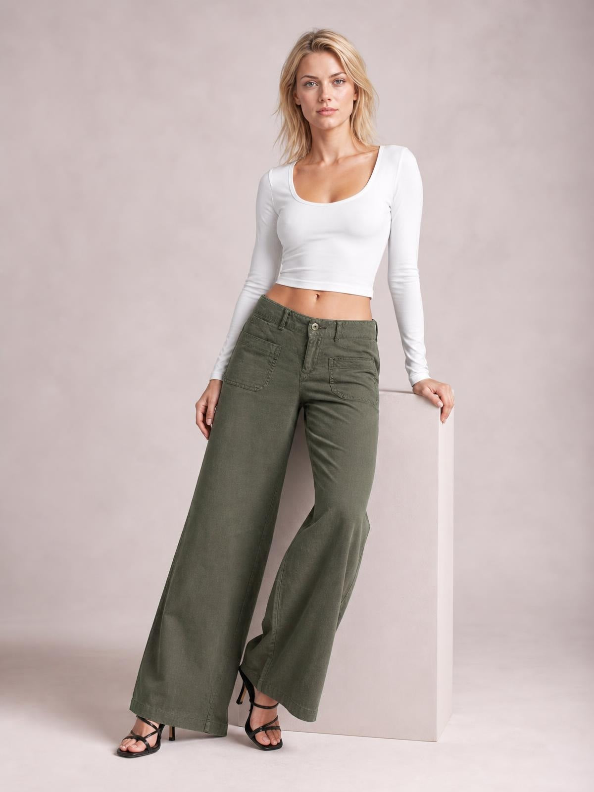 Model wears olive green wide-leg pants with front pockets and a button closure, styled with a fitted white long-sleeve top and black heeled sandals.