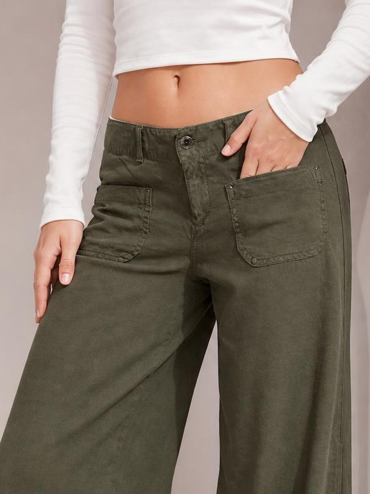 Model wearing olive green wide-leg pants with a button fly, belt loops, and multiple stitched pockets, with one hand resting in a pocket.