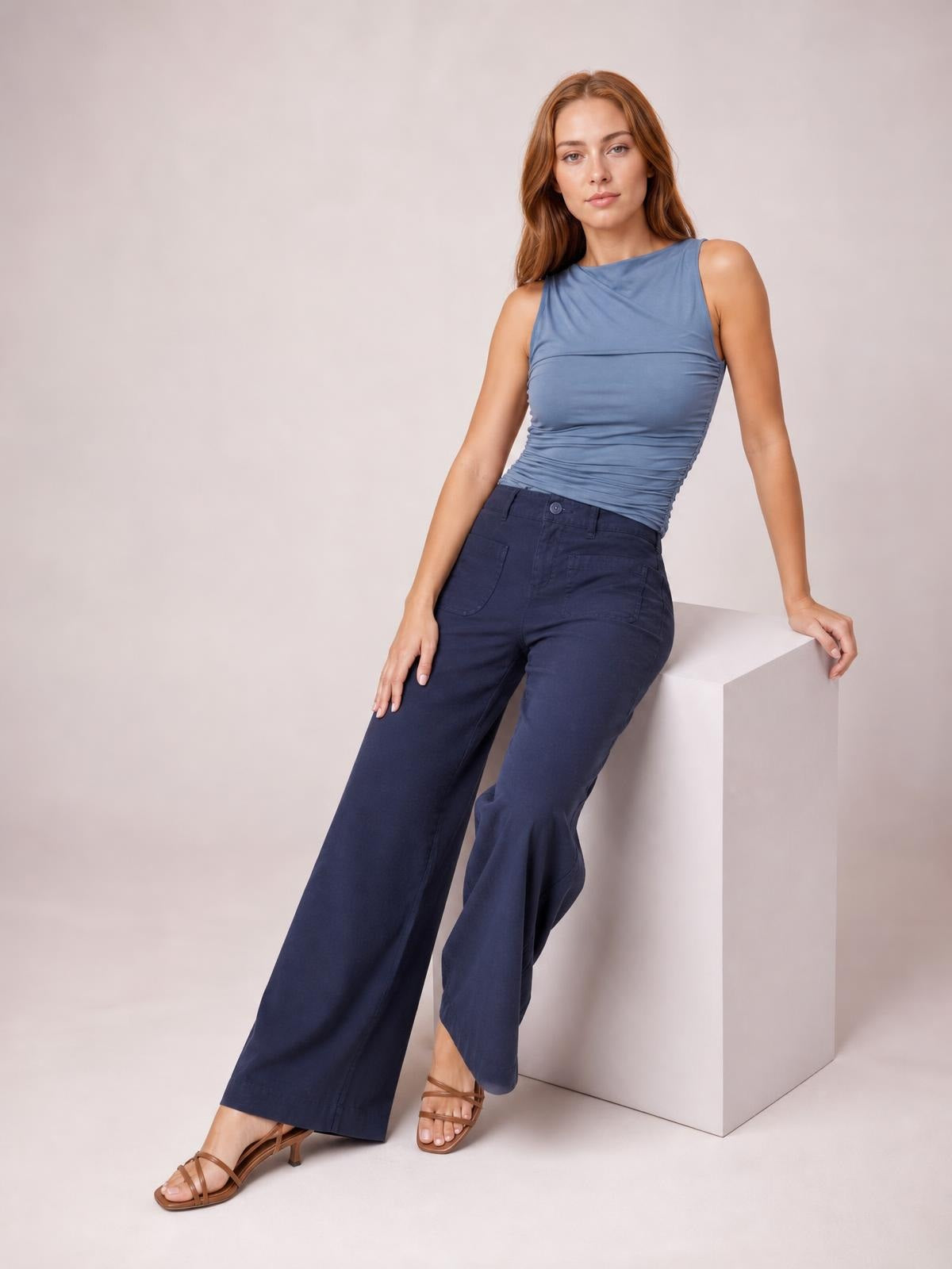 A model leans against a cube wearing navy wide-leg pants with a buttoned waistband, belt loops, curved front pockets, and a long flowing silhouette.