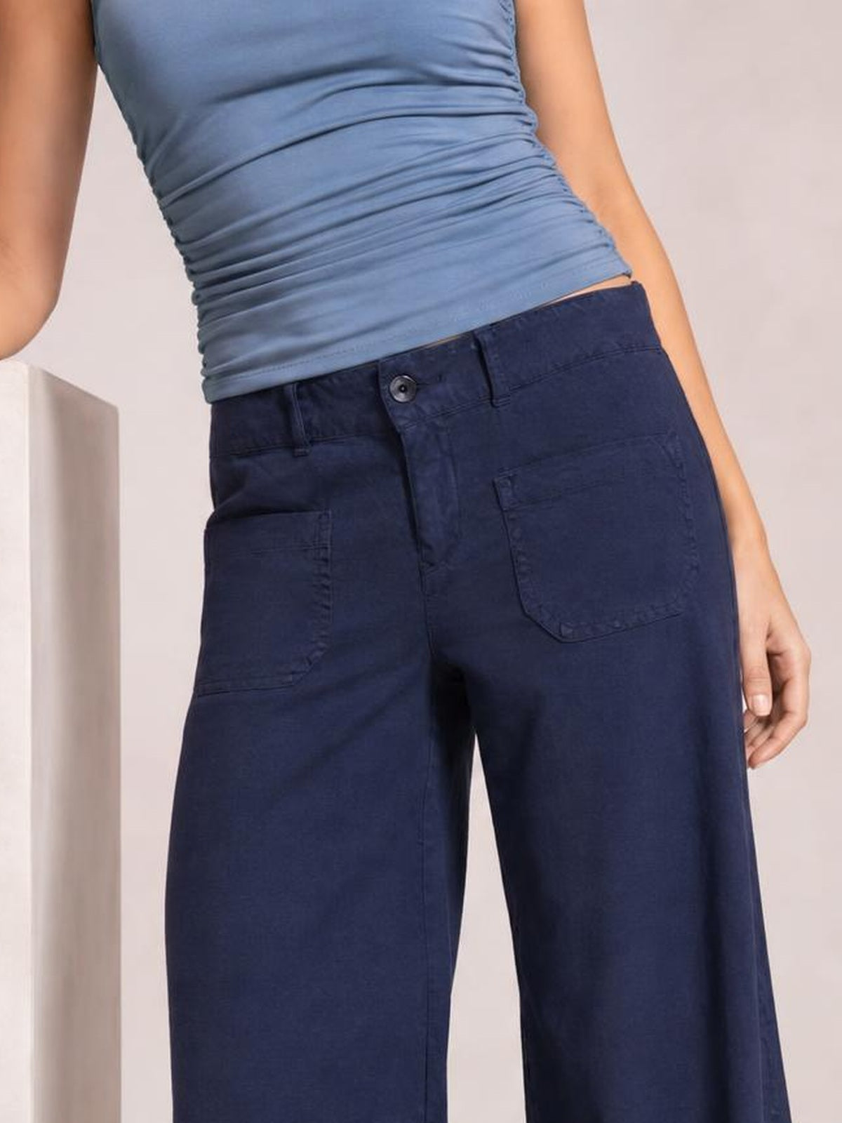 A model wears navy wide-leg pants with a buttoned waistband, belt loops, visible stitching, and roomy patch pockets in a relaxed silhouette.
