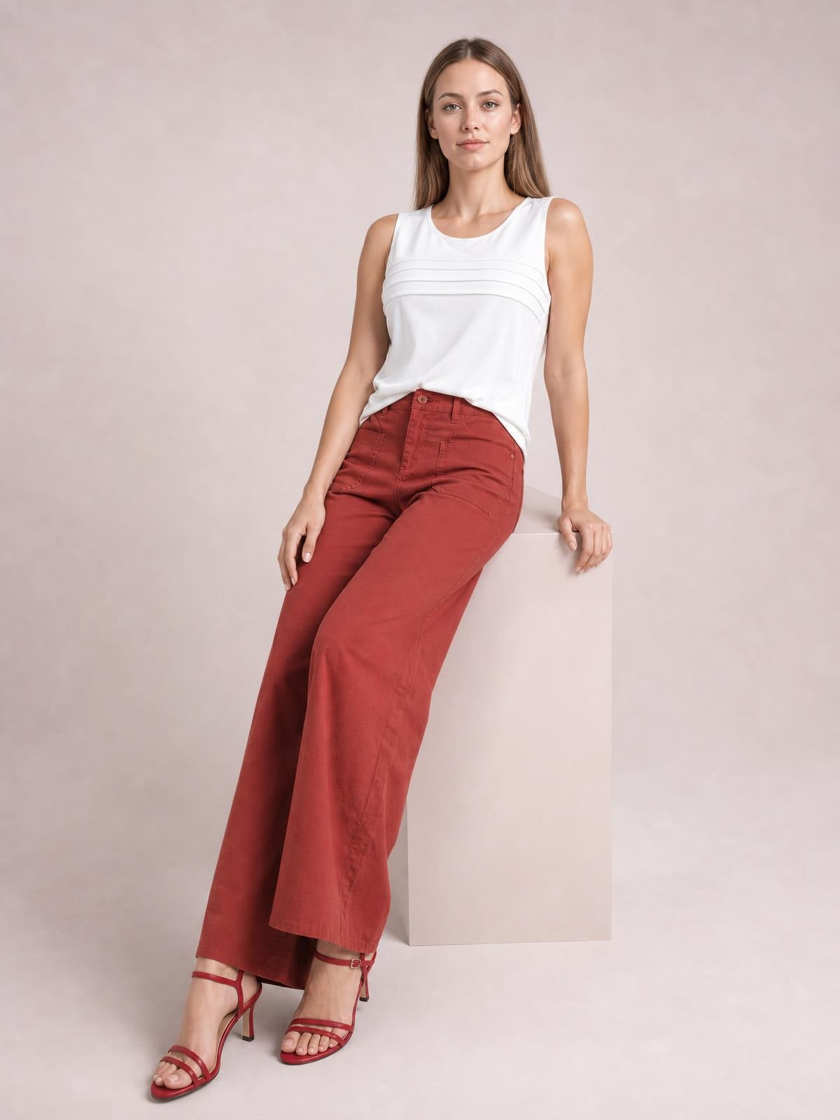 A model leans against a block wearing red wide-leg pants with a high-rise waistband, button closure, belt loops, front pockets, and a relaxed full-length silhouette.