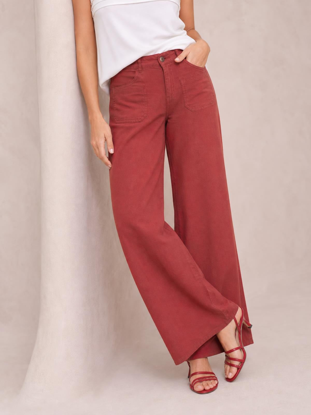 Model wears burgundy wide leg pants with a button closure, front patch pockets, and a relaxed silhouette, posing with one hand in a pocket.