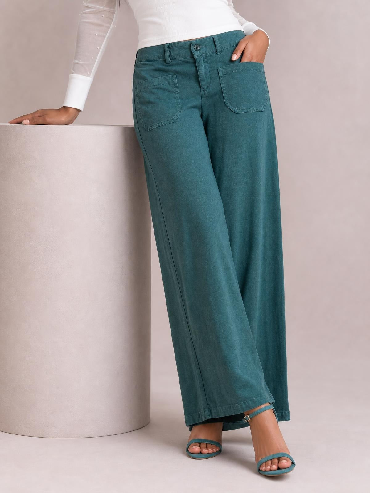 A model wears teal wide leg pants with front patch pockets, belt loops, button closure, and a relaxed full-length silhouette.