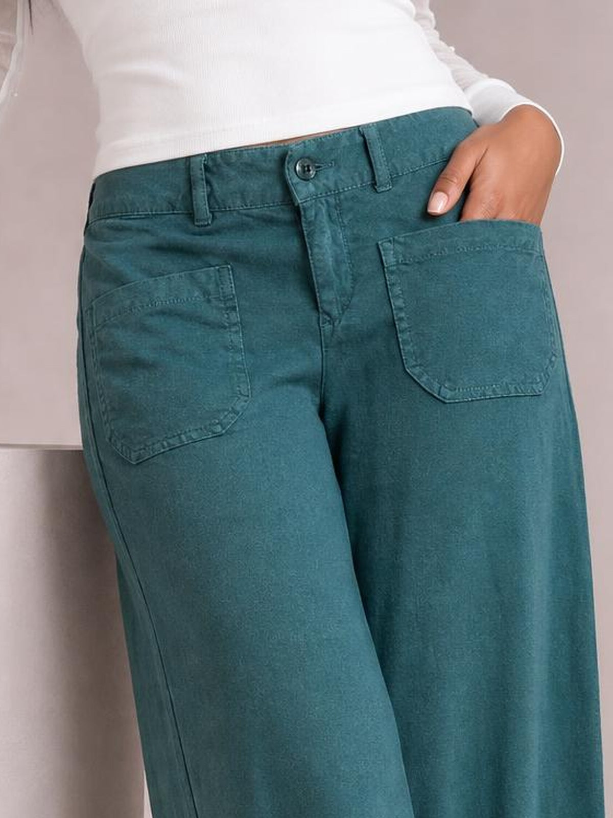 Model wears teal wide leg pants with a structured waistband, belt loops, button closure, and large stitched patch pockets, with one hand tucked inside.