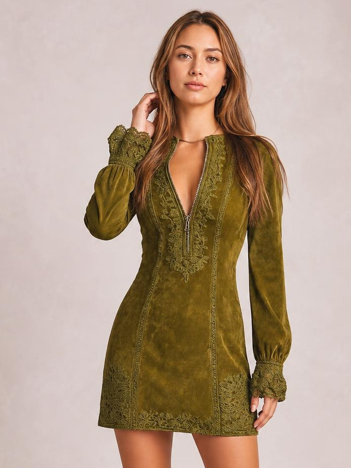 A model poses in an olive-green long-sleeve mini dress with a fitted silhouette, zip-front closure, ornate lace trim, and gathered cuffs.