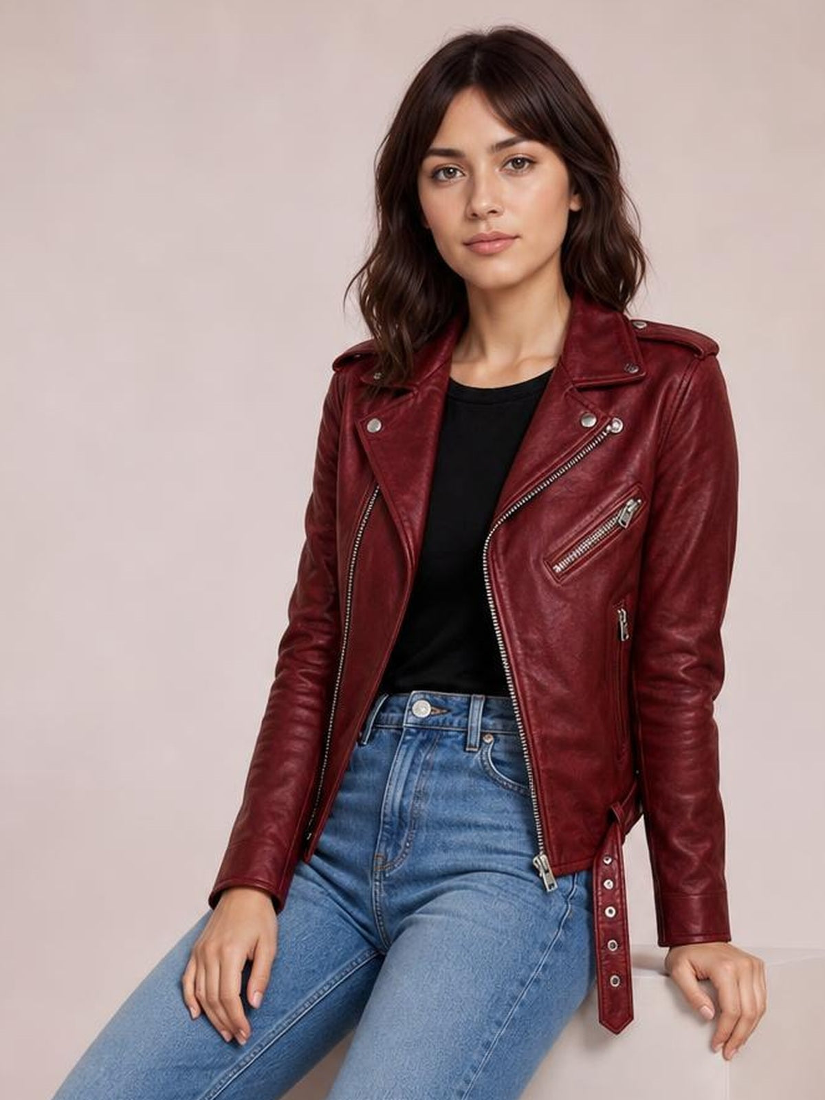 A seated model wears a burgundy leather-look motorcycle biker jacket with an asymmetric zip front, belted waist, zippered pockets, and fitted sleeves.