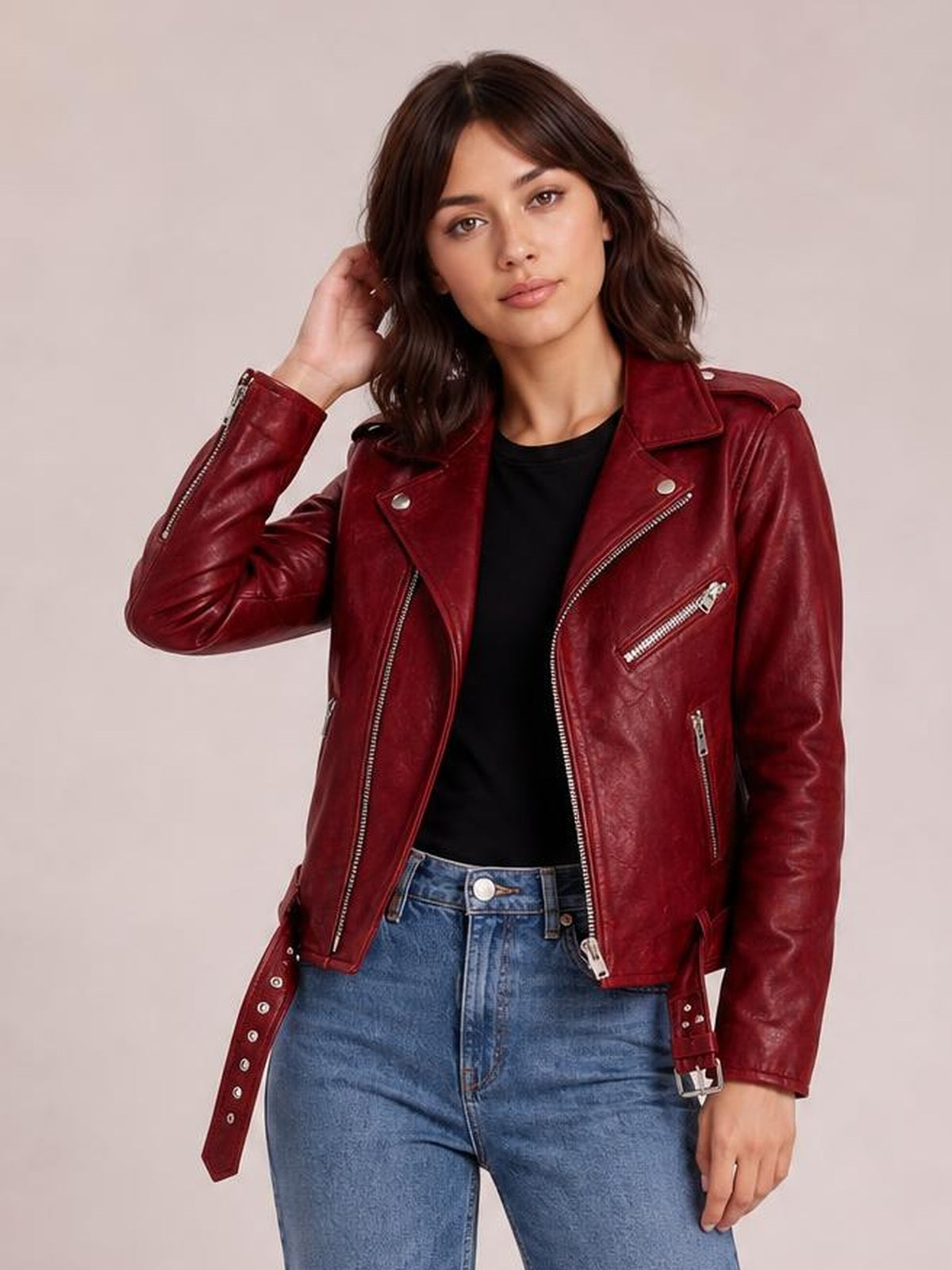 Model poses in a burgundy leather motorcycle biker jacket with an asymmetrical zip, wide lapels, zippered pockets, belted cuffs, and a cropped fitted silhouette.