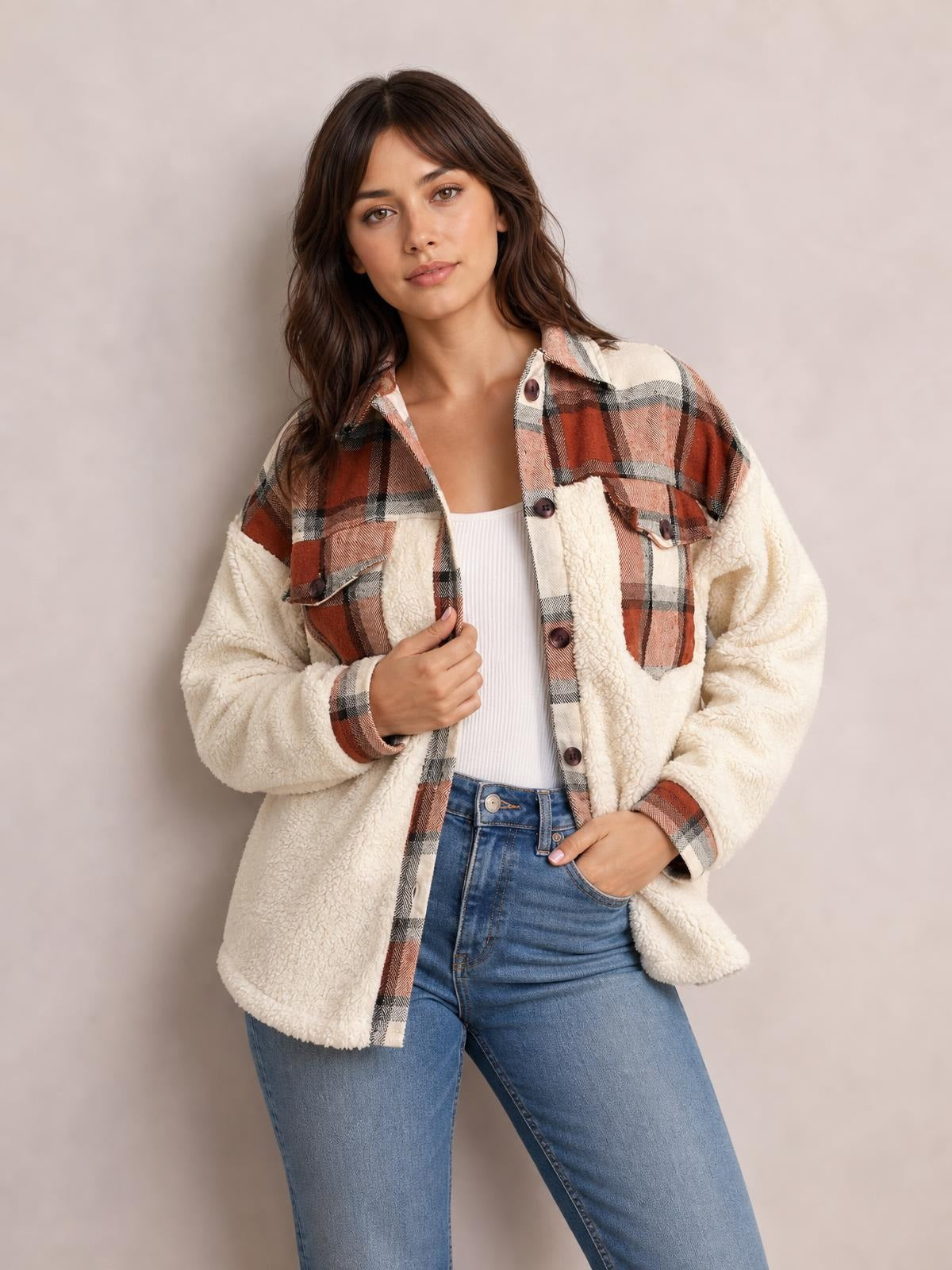 Model wears a fuzzy cream button-up jacket with long sleeves, rust plaid panels, flap chest pockets, and a relaxed silhouette while holding the front.