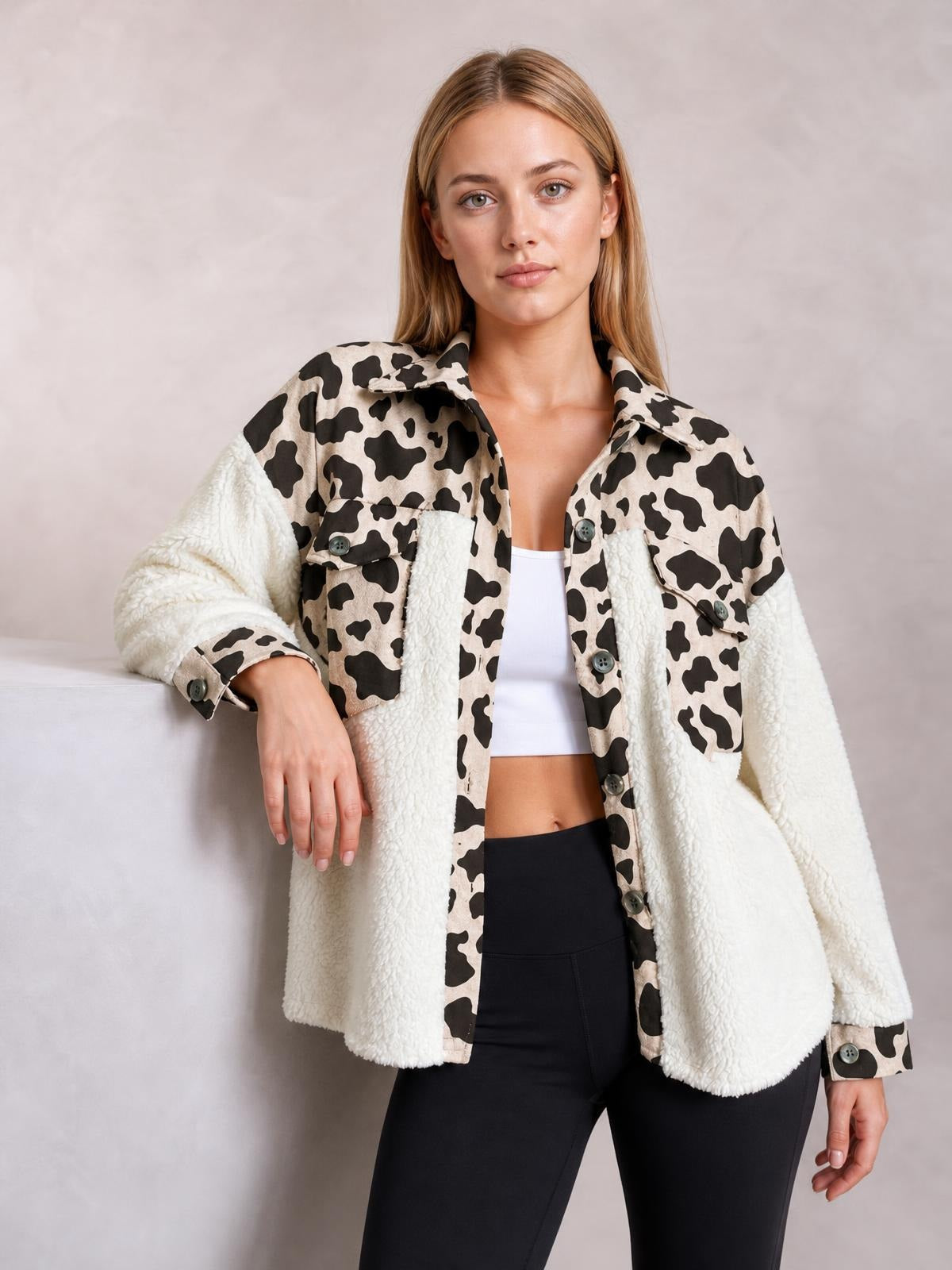 A model wears a cream-and-black cow-print fuzzy button-up jacket with white sherpa panels, long sleeves, chest pockets, and a relaxed silhouette while leaning on one arm.