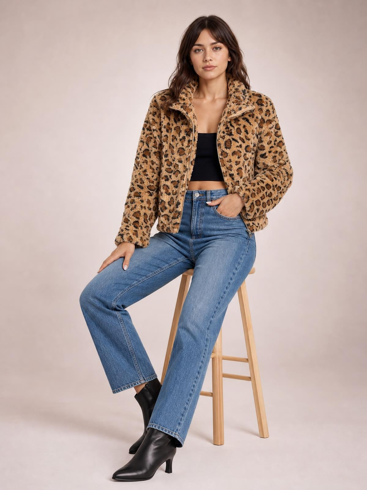 A model sits with one hand in her jeans pocket, wearing a fuzzy tan leopard zip-up jacket with a high turtleneck collar and relaxed silhouette.