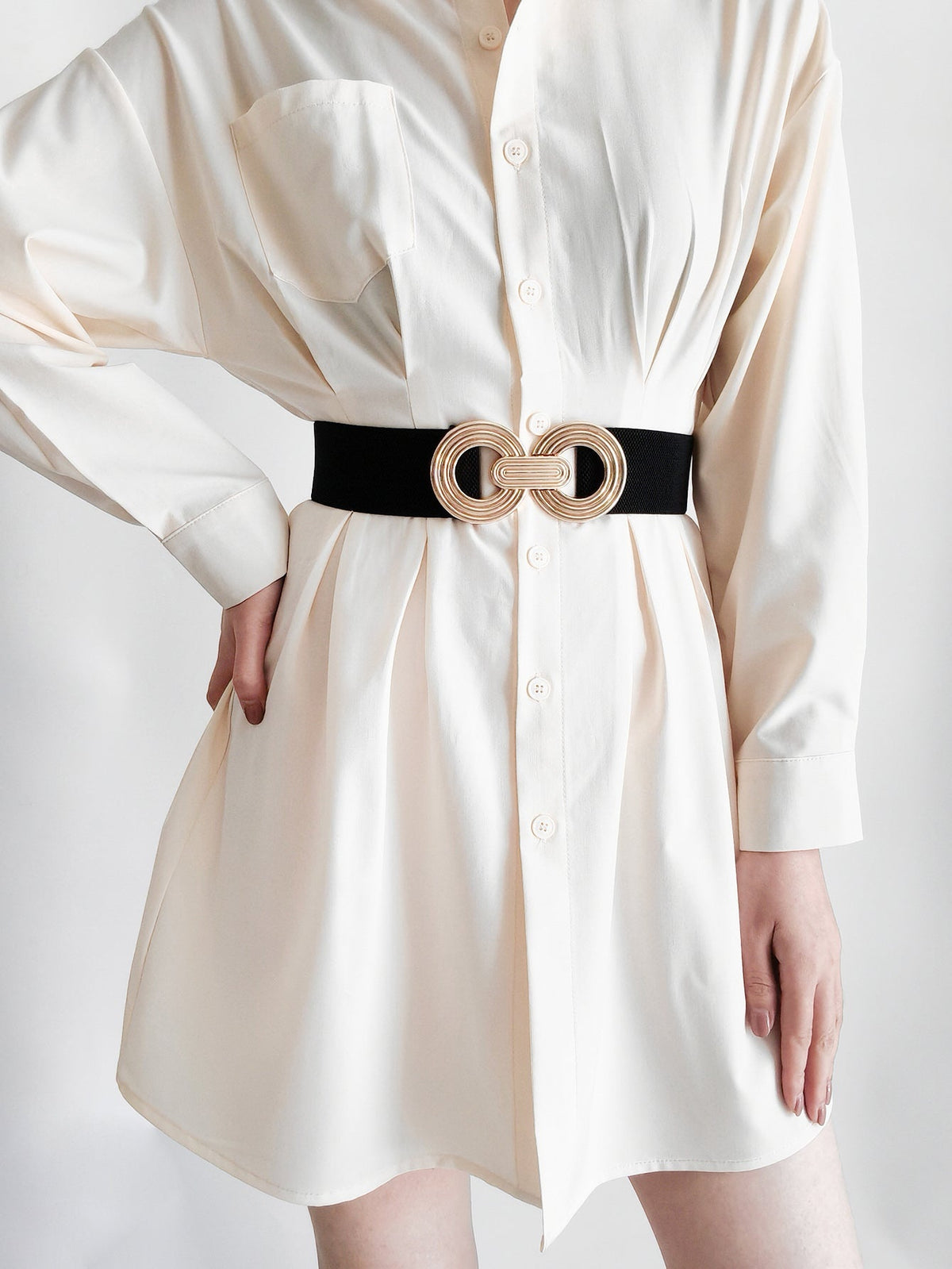 Model in a white shirt dress wears a black elastic wide belt with a sculptural gold geometric buckle, defining the waist in a relaxed silhouette.