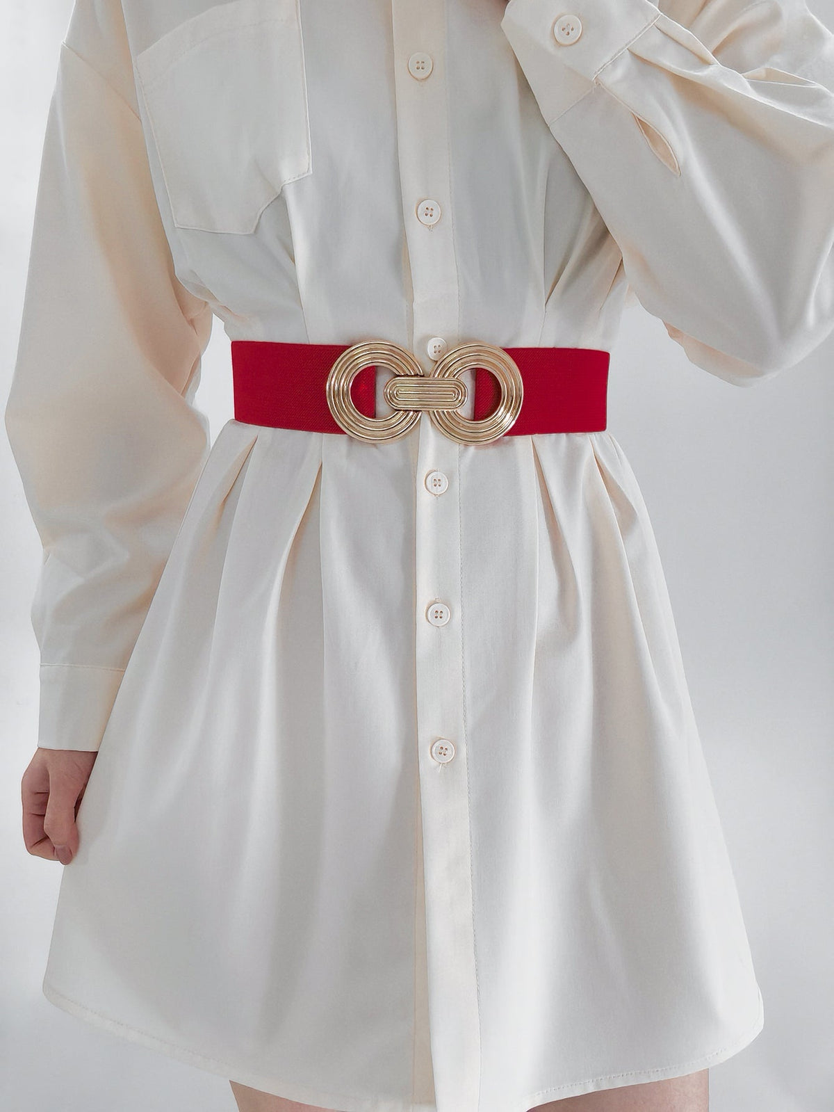 A model wears a red elastic wide belt over a white button-front shirt dress, featuring a sculptural geometric alloy buckle that defines the waist.