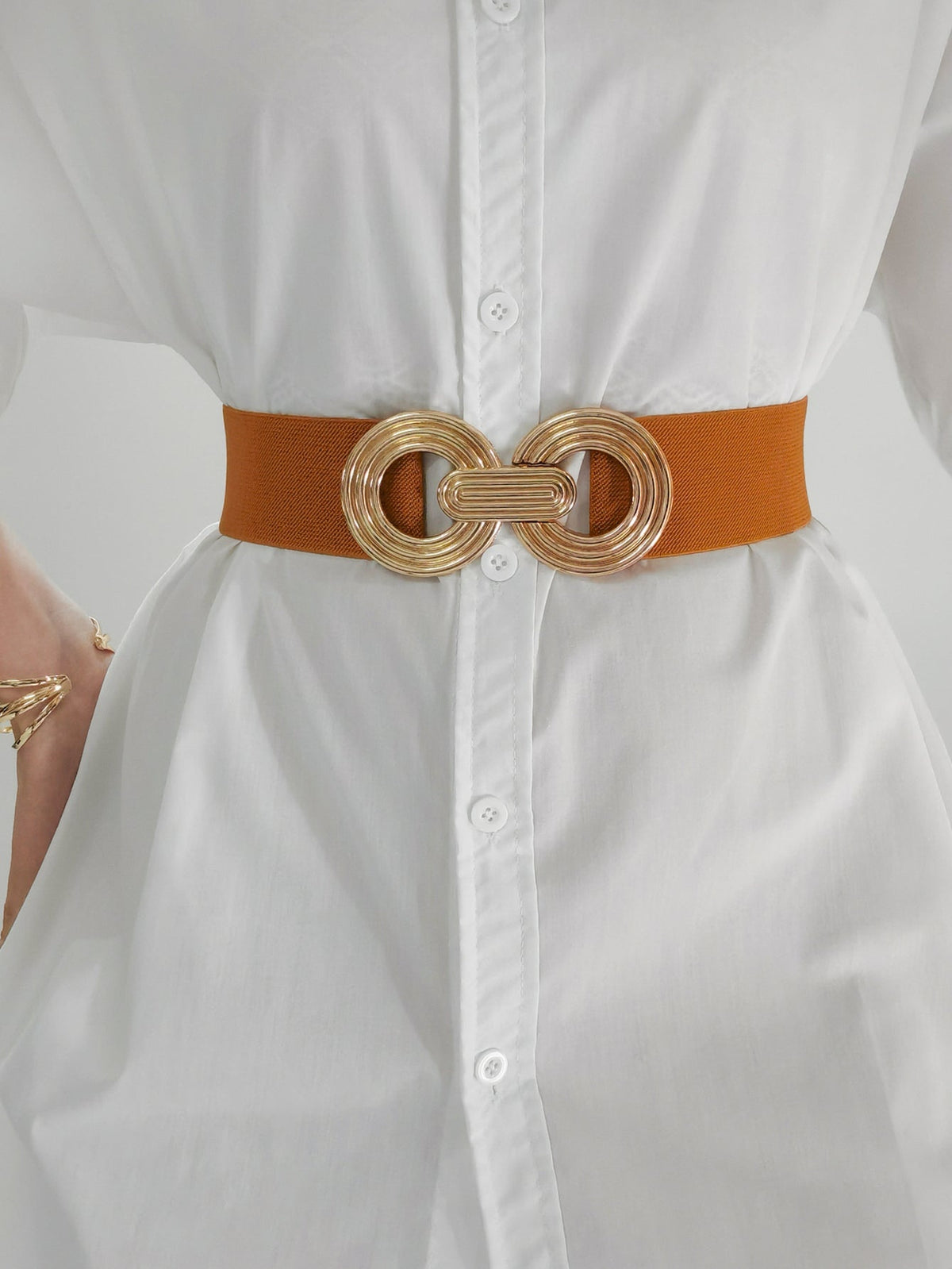 Close-up of a model wearing a brown elastic wide belt over a white button-front shirt, featuring a sculptural gold geometric buckle and defined waist silhouette.