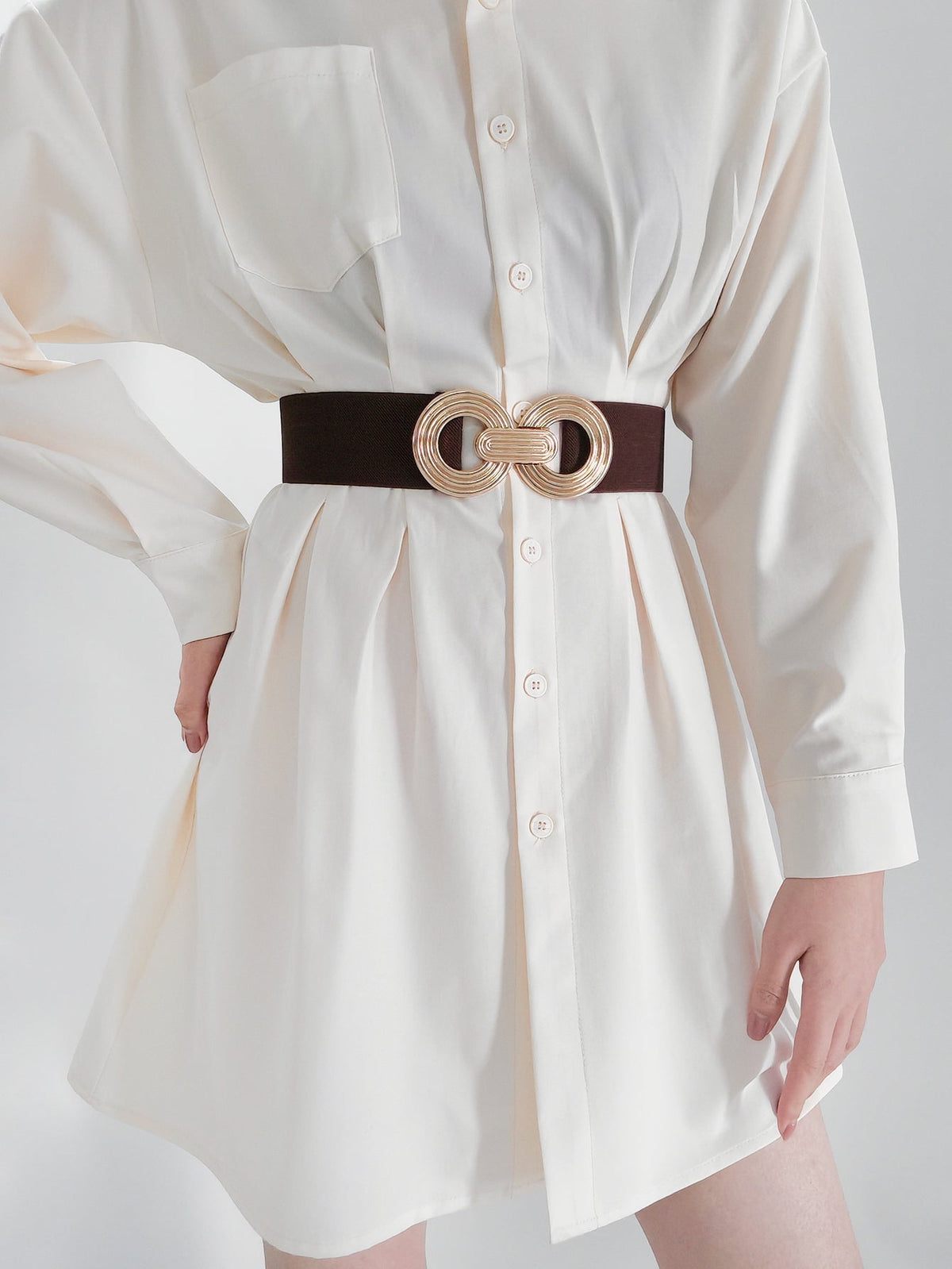 A model wears a white button-front shirt dress with a defined waist, styled with a dark elastic wide belt and dimensional gold geometric buckle.