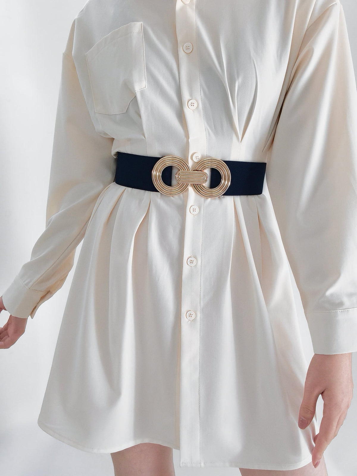 A navy elastic wide belt cinches a white shirt dress at the waist, featuring a sculptural gold geometric buckle with interlocking rounded forms.