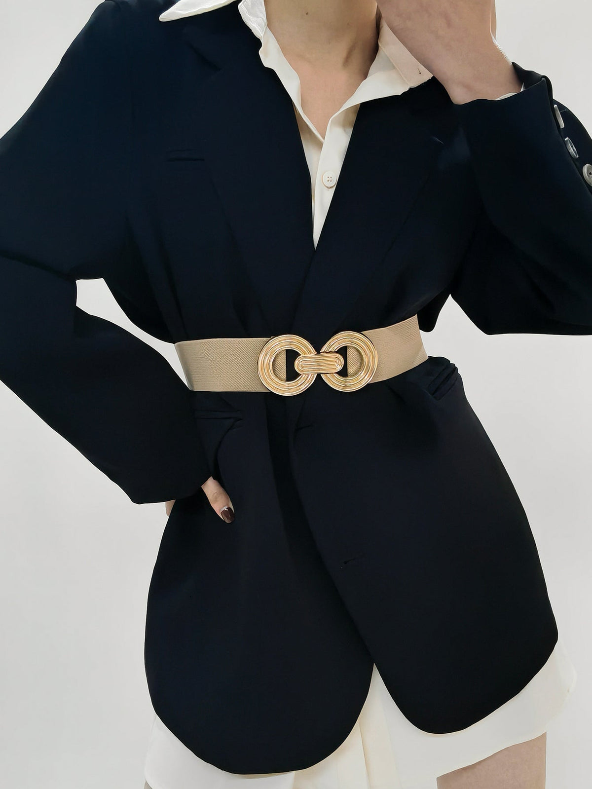 Model poses with one hand on her hip, wearing a beige elastic wide belt over a black blazer, finished with a dimensional gold geometric buckle.