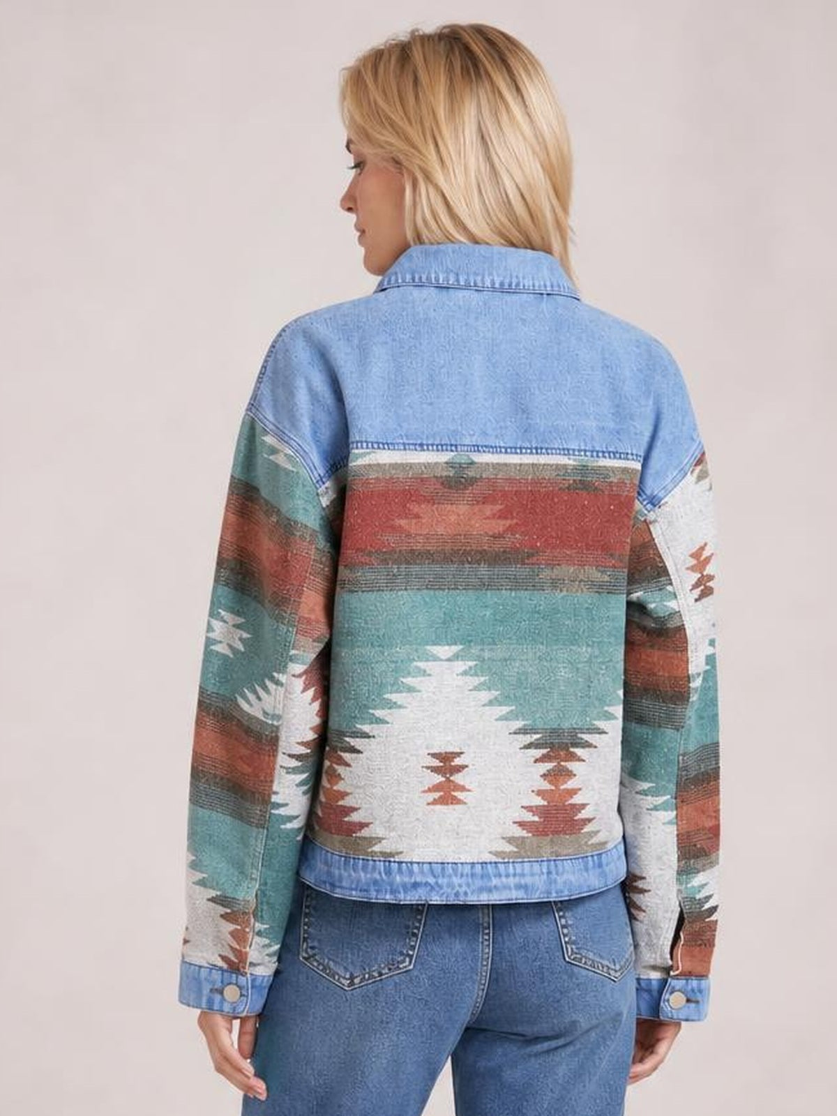 A model stands turned away in a light blue denim jacket with dropped shoulders, buttoned cuffs, and colorful geometric patterns across the sleeves and back.