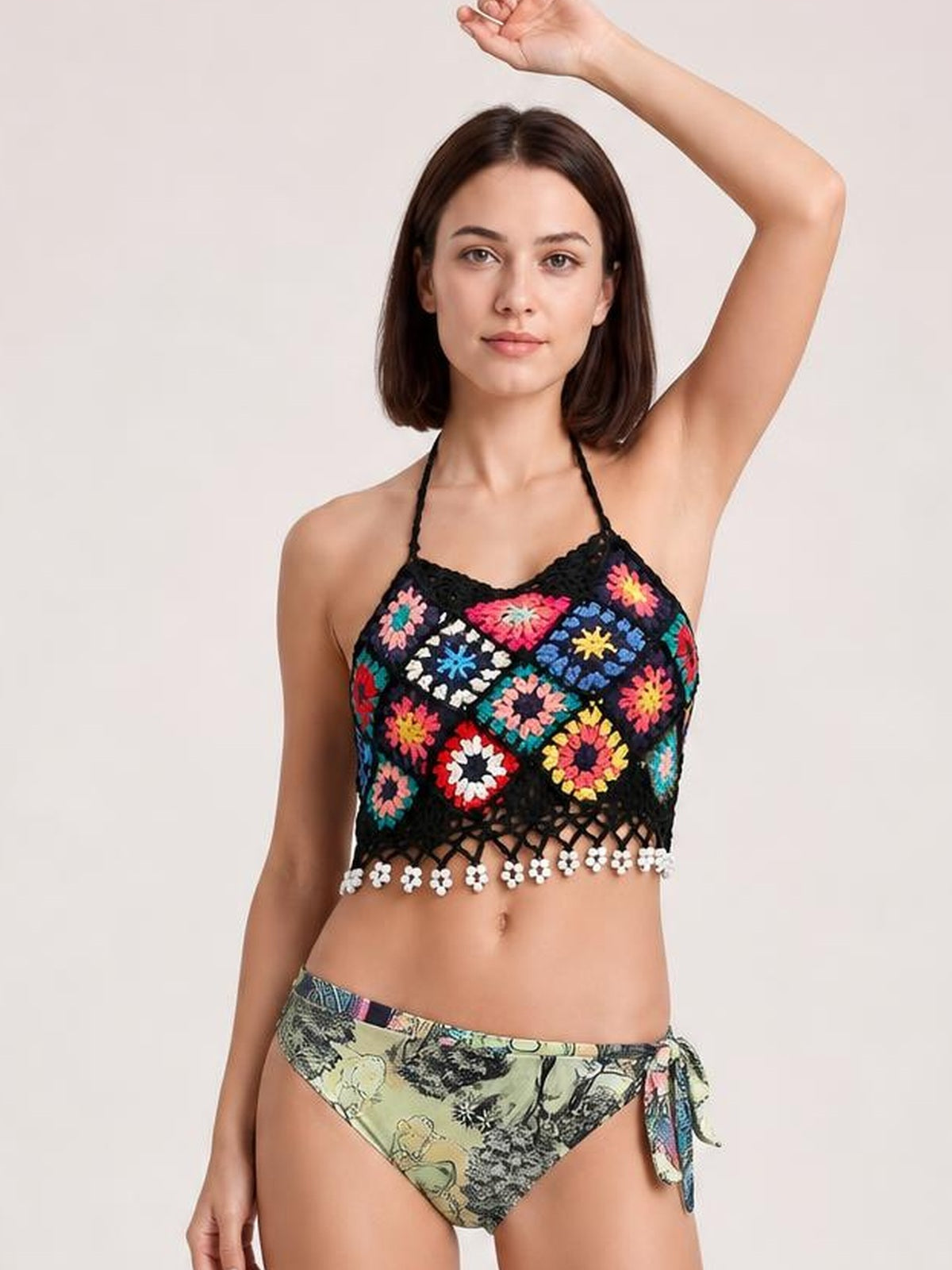 Model wearing a black geometric halter neck swim top with colorful floral crochet panels, an openwork cropped silhouette, and a decorative fringe hem.