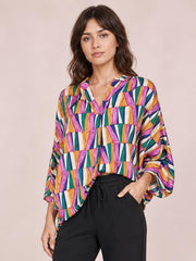 Model wearing a colorful geometric print top with a notched neckline, relaxed dolman sleeves, and a loose silhouette, posing with one hand in her pocket.