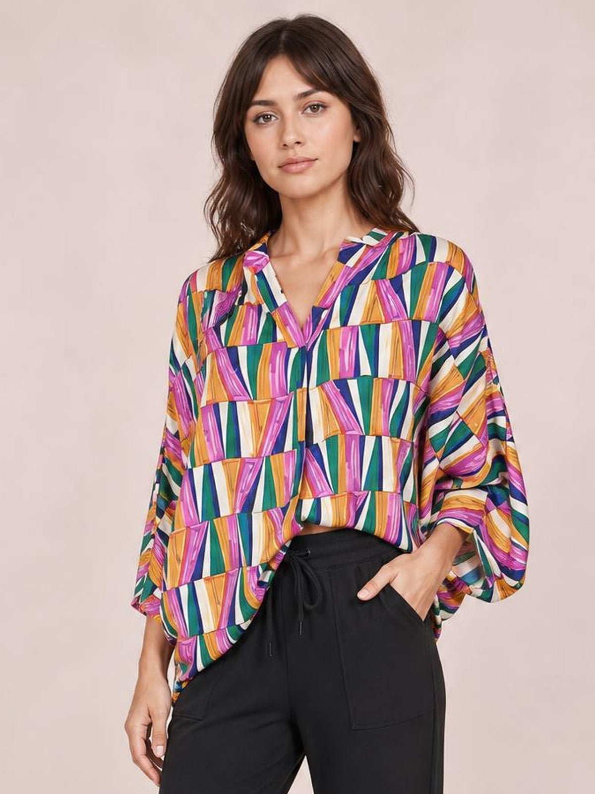 Model wearing a colorful geometric print top with a notched neckline, relaxed dolman sleeves, and a loose silhouette, posing with one hand in her pocket.