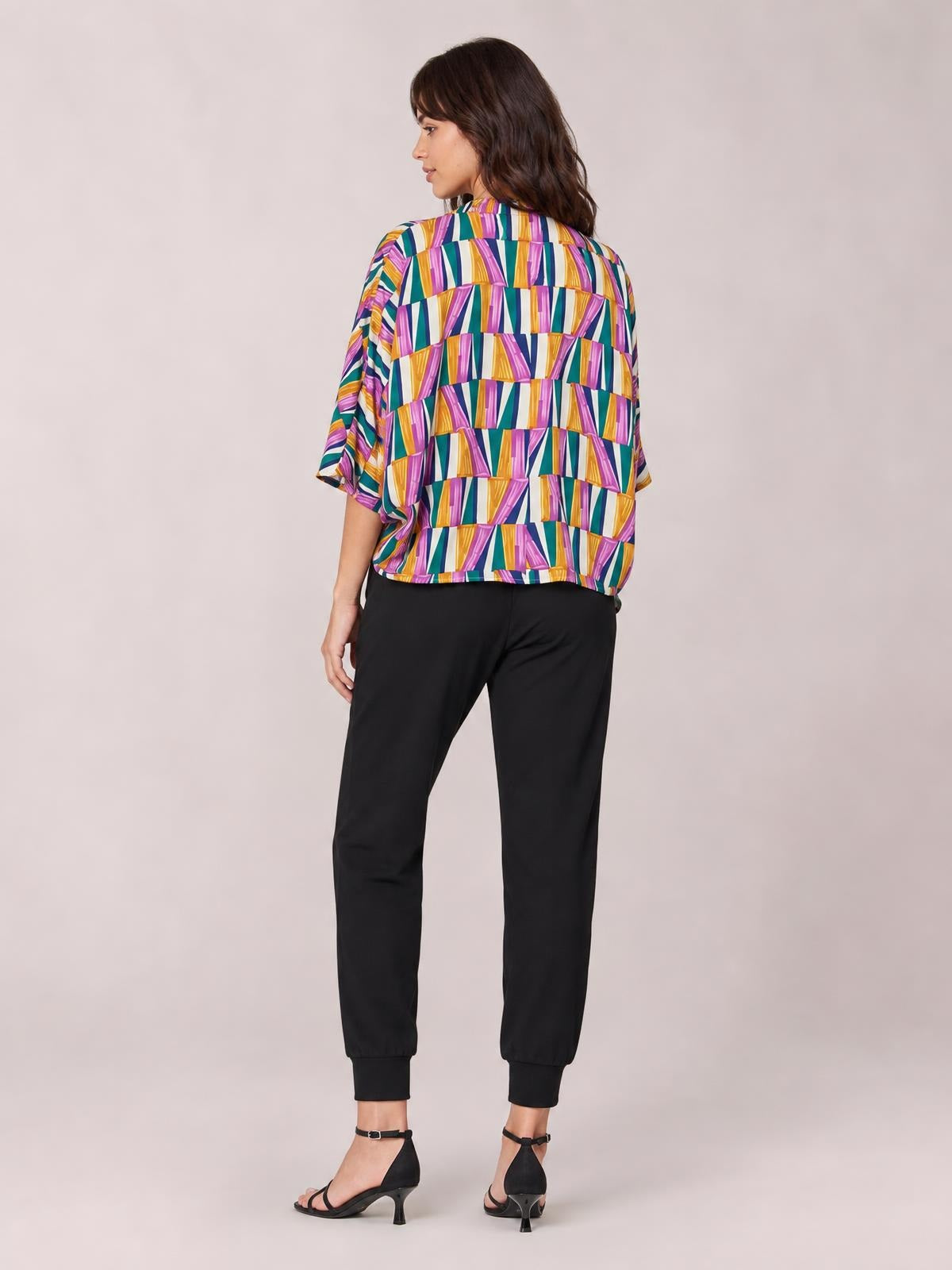 A model turns to display a colorful geometric top with loose dolman sleeves, a relaxed boxy silhouette, and a straight hem in vibrant purple, teal, yellow, and white.