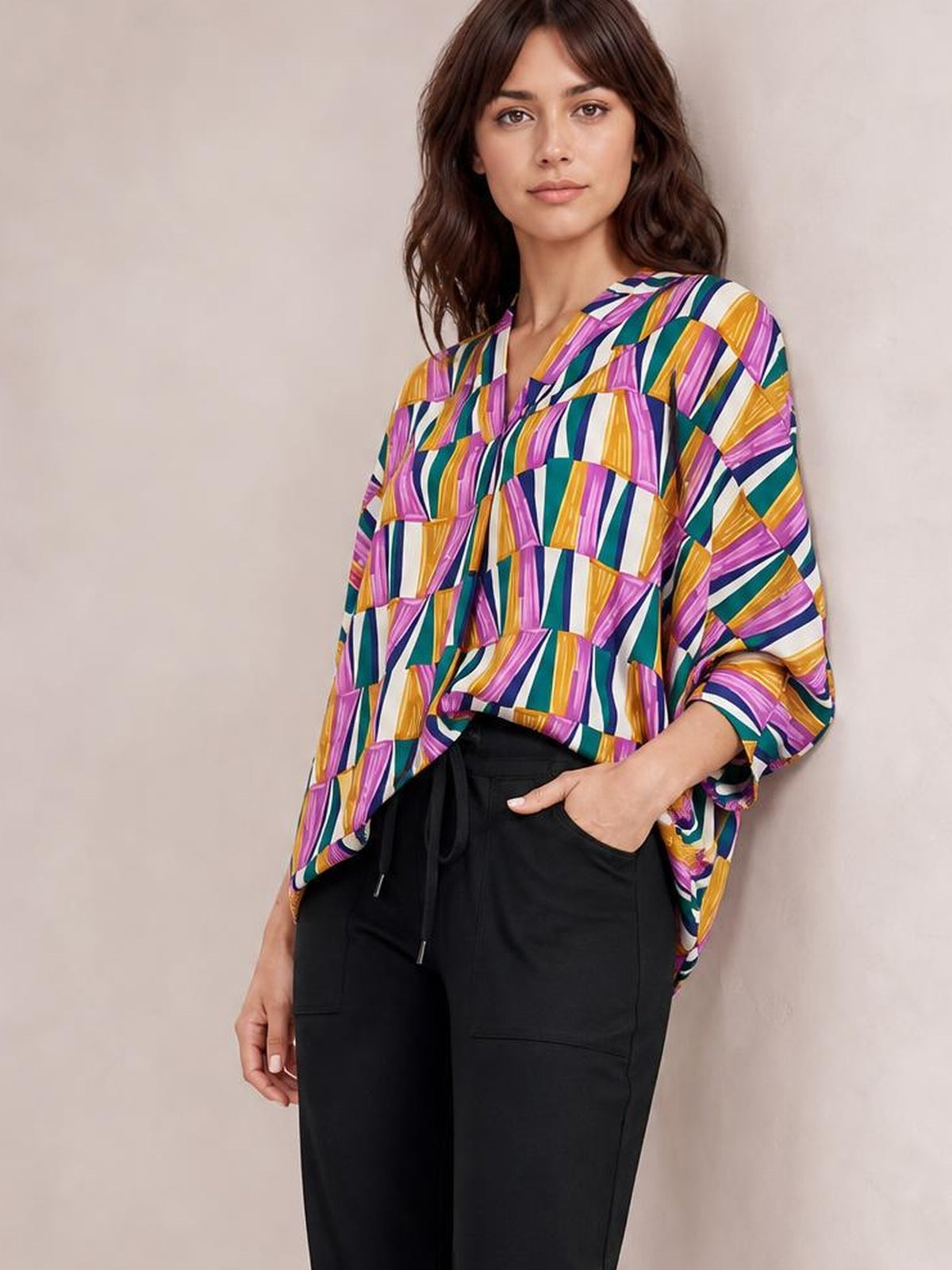 A model stands wearing a loose geometric top in purple, teal, yellow, and white with a notched neckline and relaxed dolman sleeves.