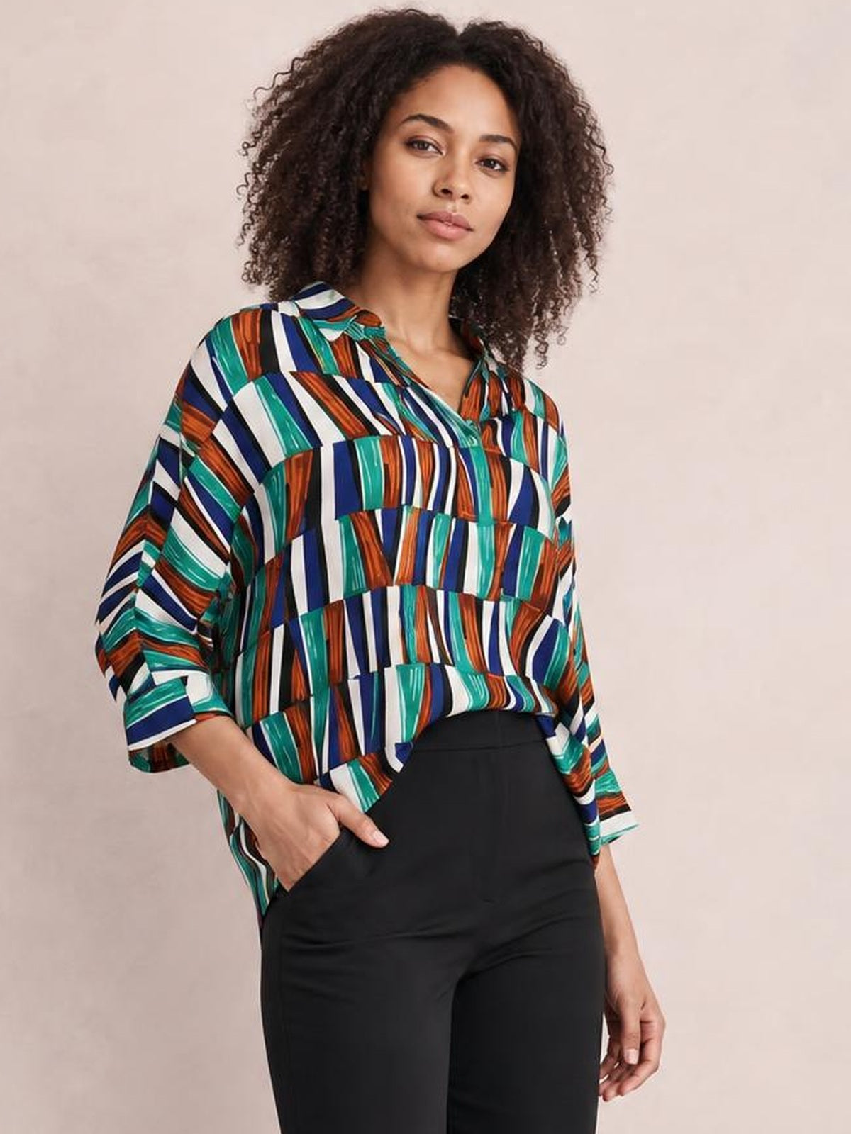 A model poses with one hand in her pocket wearing a colorful geometric notched dolman sleeve top with a relaxed silhouette and three-quarter sleeves.