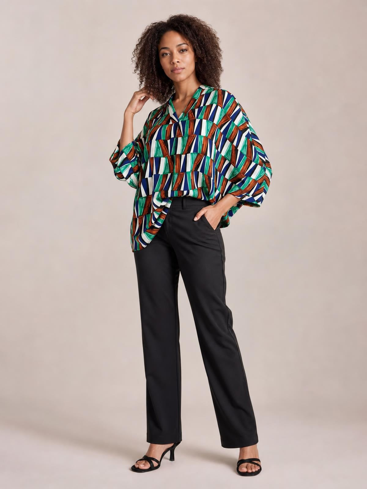 Standing model wears a multicolor geometric top with a notched neckline, loose dolman sleeves, and a relaxed silhouette in green, blue, orange, white, and black.