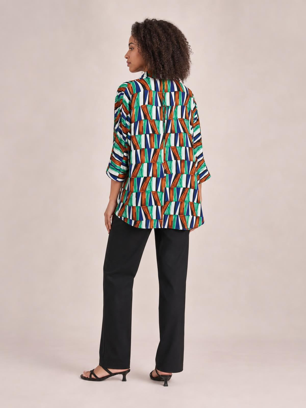 A model stands in profile-back view wearing a loose geometric top with colorful navy, green, orange, and white print, dolman sleeves, and a curved hem.