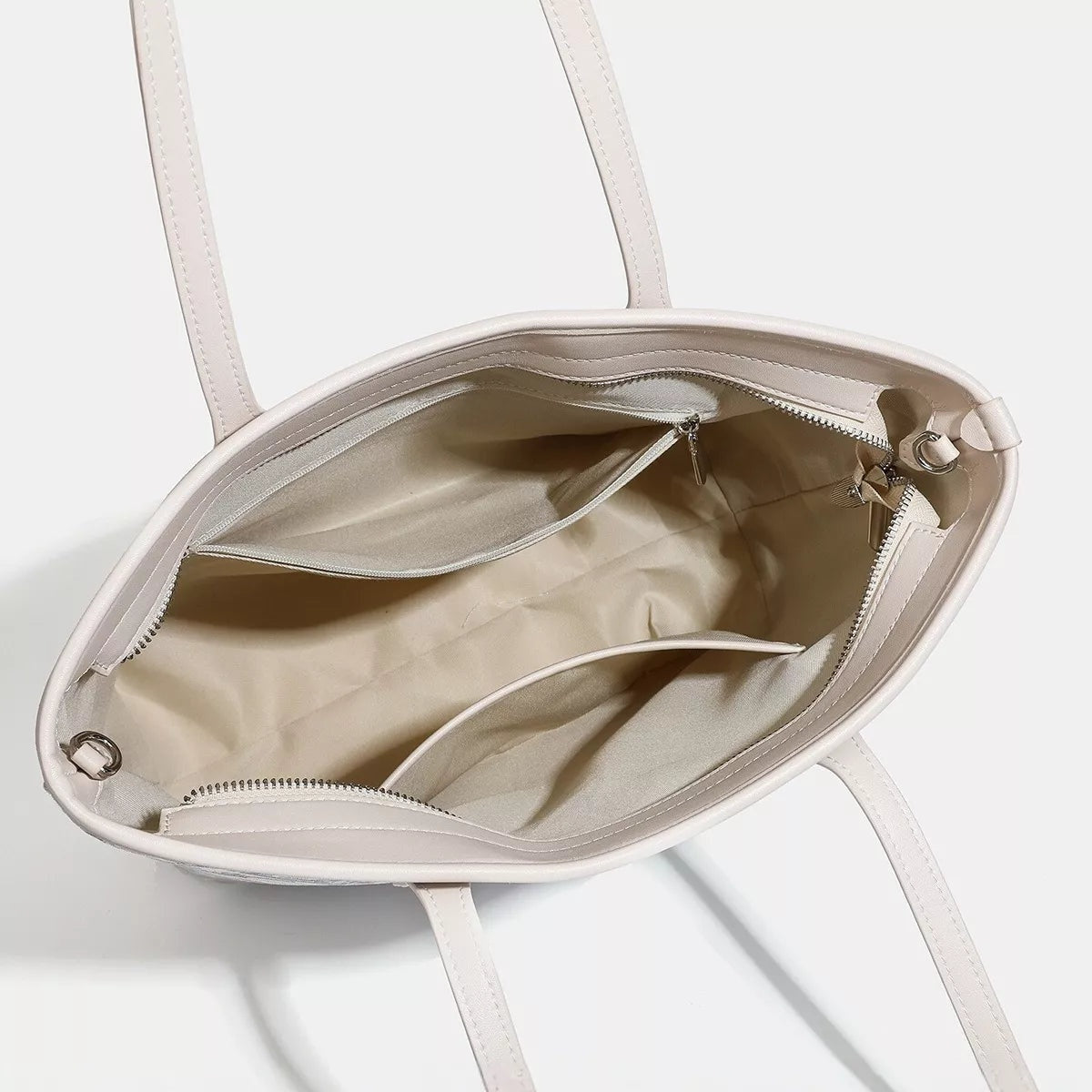 Open medium faux leather tote bag in a light neutral color, showing long handles, a zippered top, spacious lining, and interior compartments.