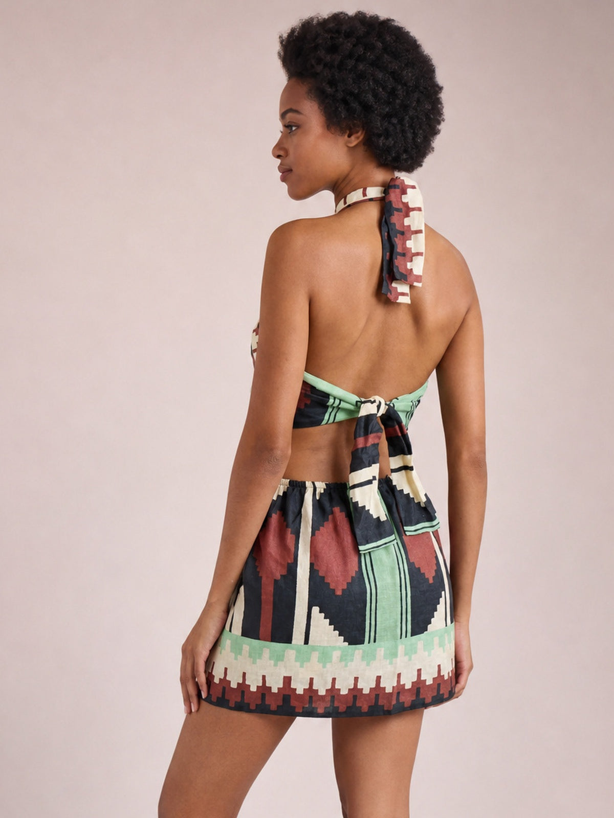 A model shows the back of a geometric print halter neck mini dress in sage, black, red, and cream with a backless tied design and short A-line silhouette.