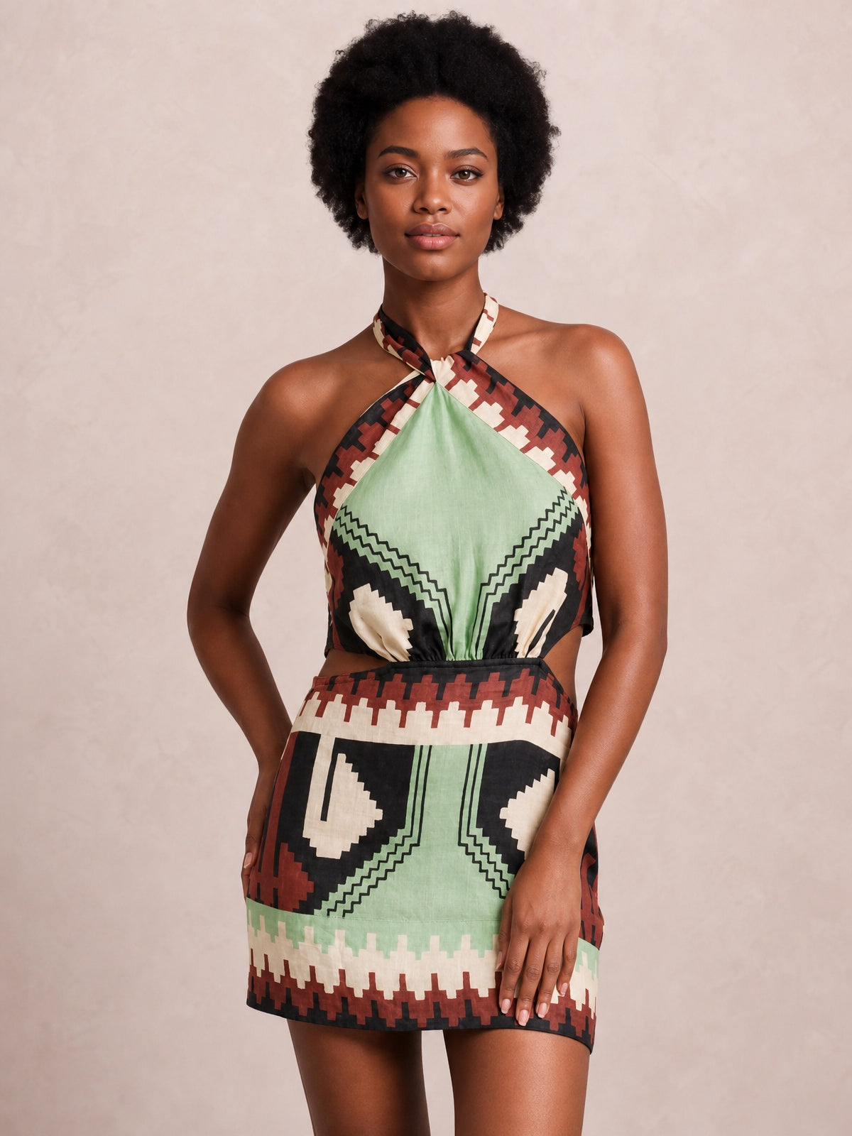 Model poses with one hand in her pocket wearing a sage-led geometric print halter neck mini dress with a cutout waist and short, structured silhouette.