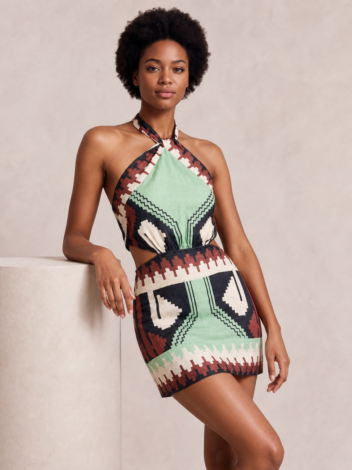 Model poses in a geometric print halter neck mini dress with a sage-led pattern, bold contrasting shapes, a waist cutout, and a streamlined short silhouette.