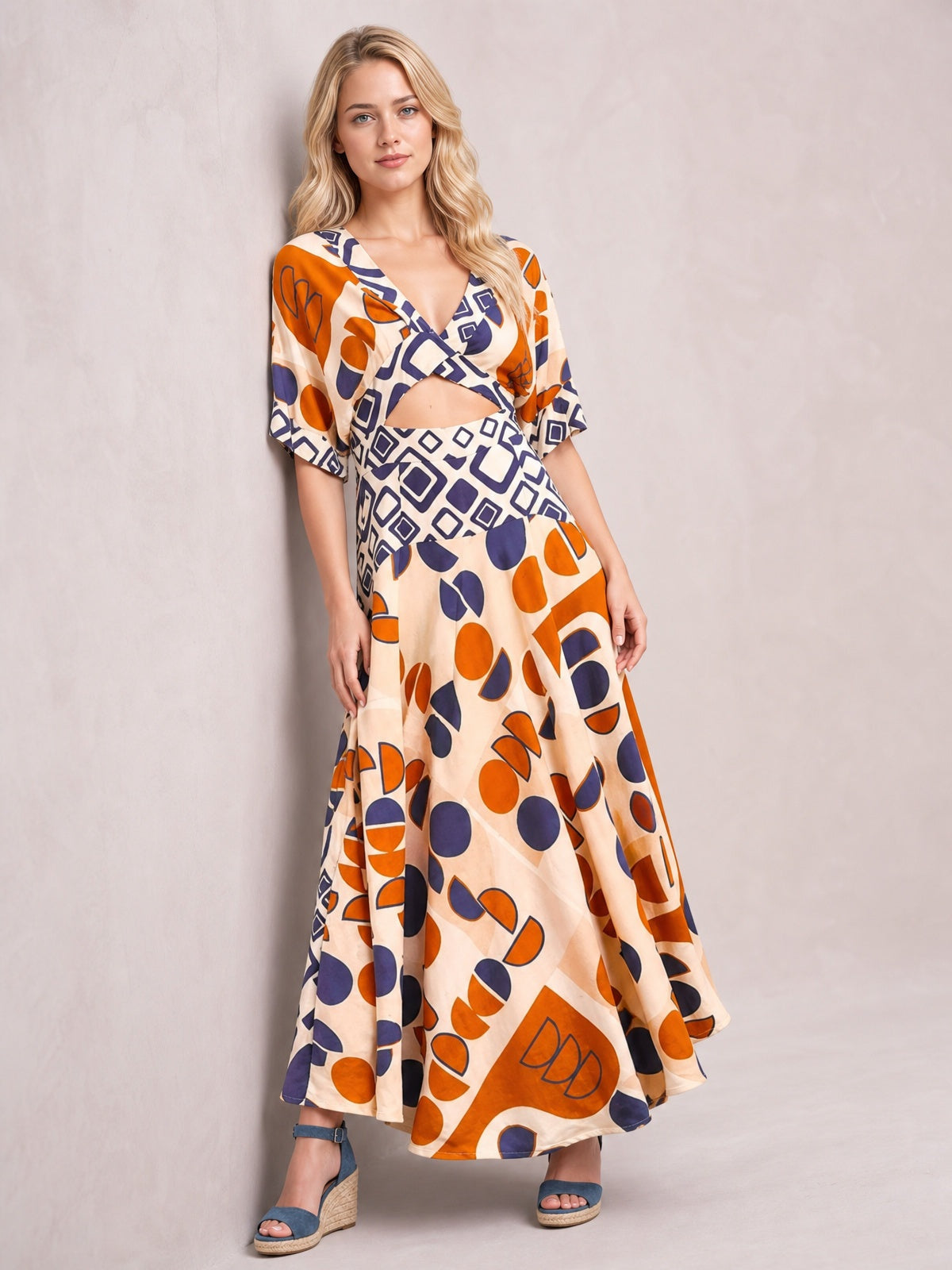 Model poses in a geometric print V-neck maxi dress with a caramel base, navy and orange motifs, short sleeves, waist cutout, and flowing floor-length silhouette.