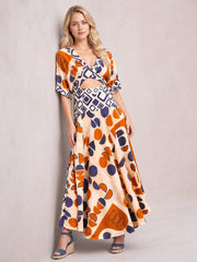 Model poses in a geometric print V-neck maxi dress with a caramel base, navy and orange motifs, short sleeves, waist cutout, and flowing floor-length silhouette.