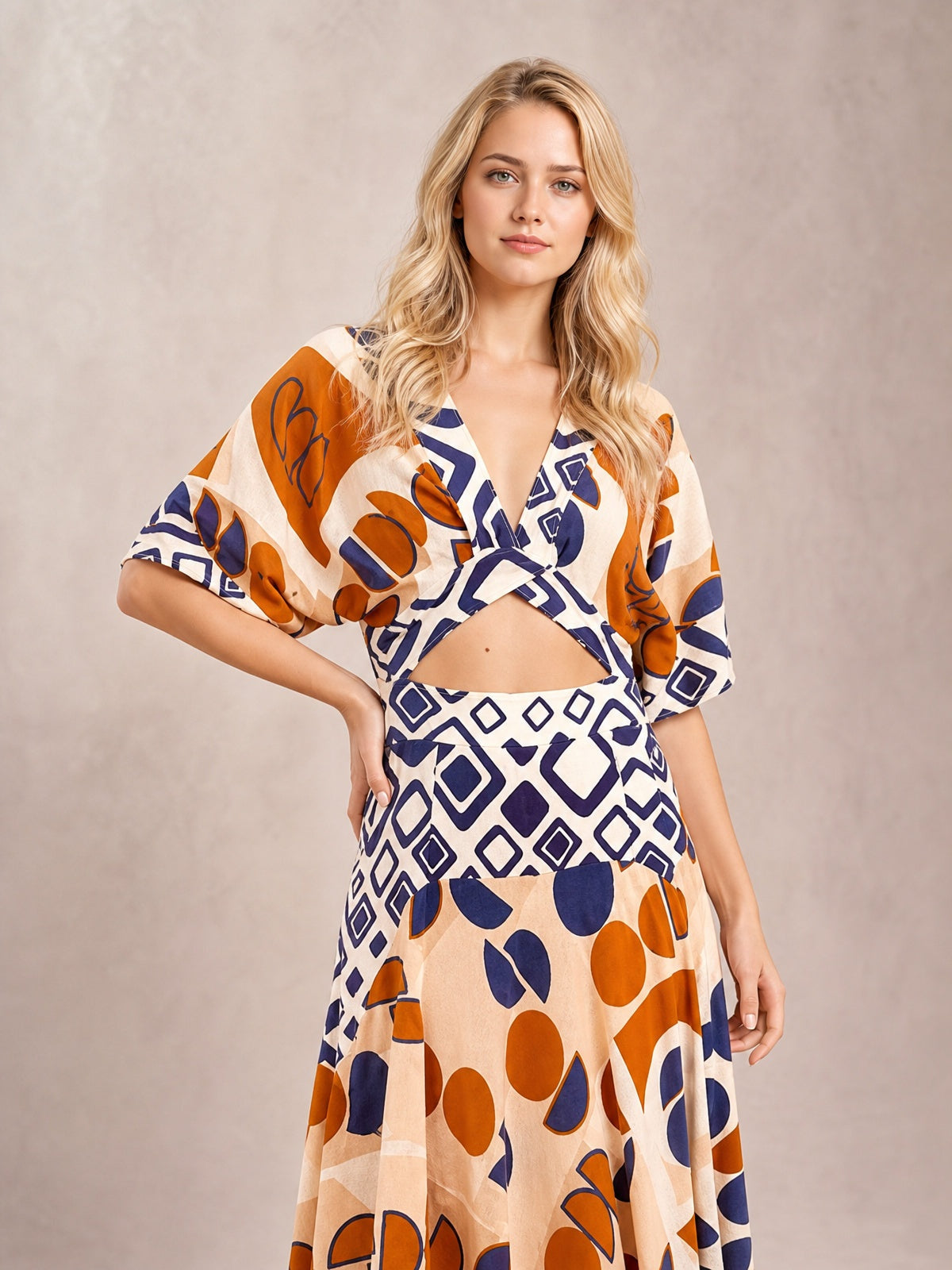Model poses with one hand on her hip in a geometric print maxi dress featuring a V-neck, waist cutout, short sleeves, and a flowing caramel, navy, orange, and cream silhouette.