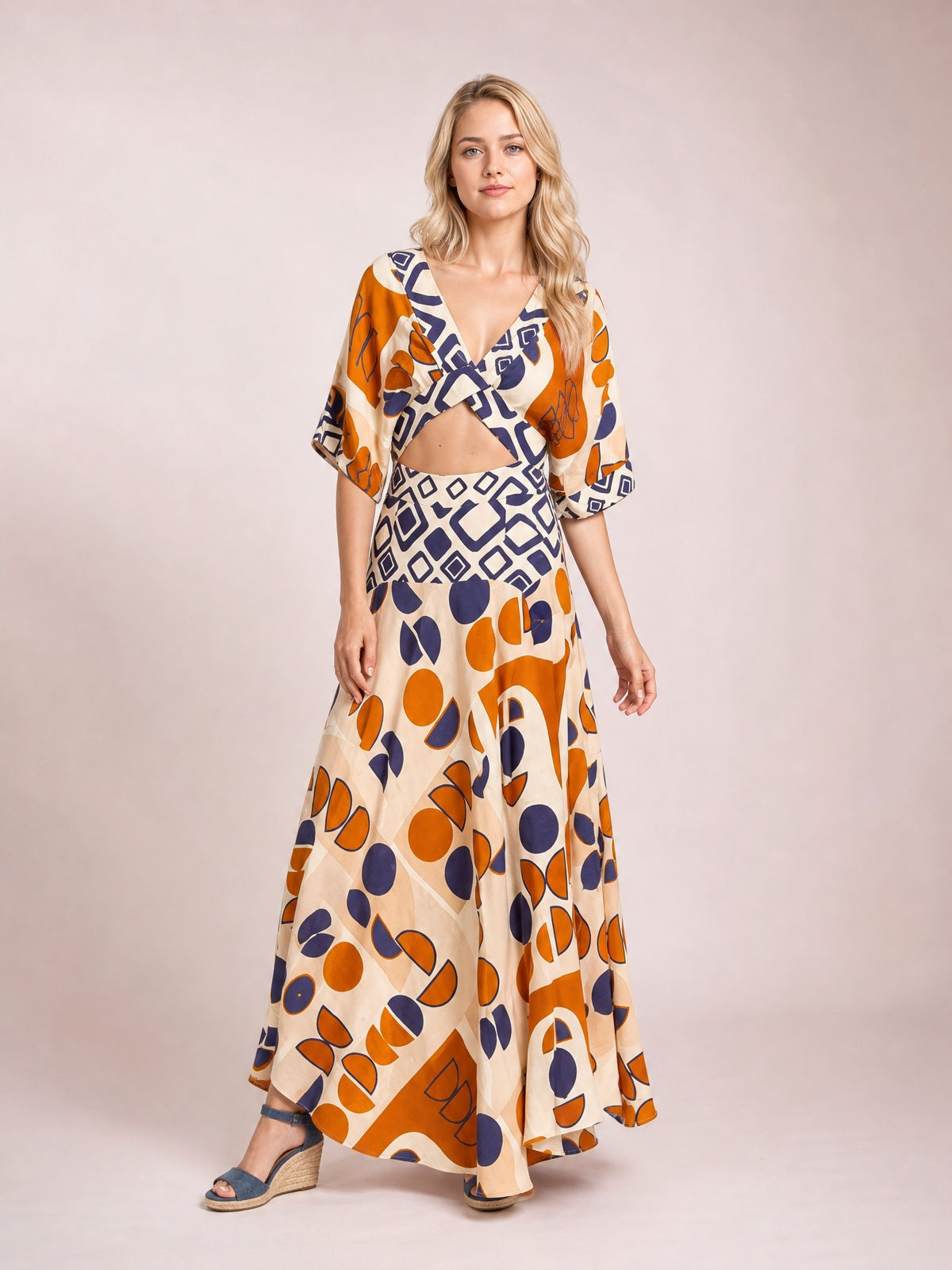 A model stands in a geometric print maxi dress with a caramel and cream base, navy and orange motifs, V-neck, waist cutout, short sleeves, and flowing floor-length skirt.