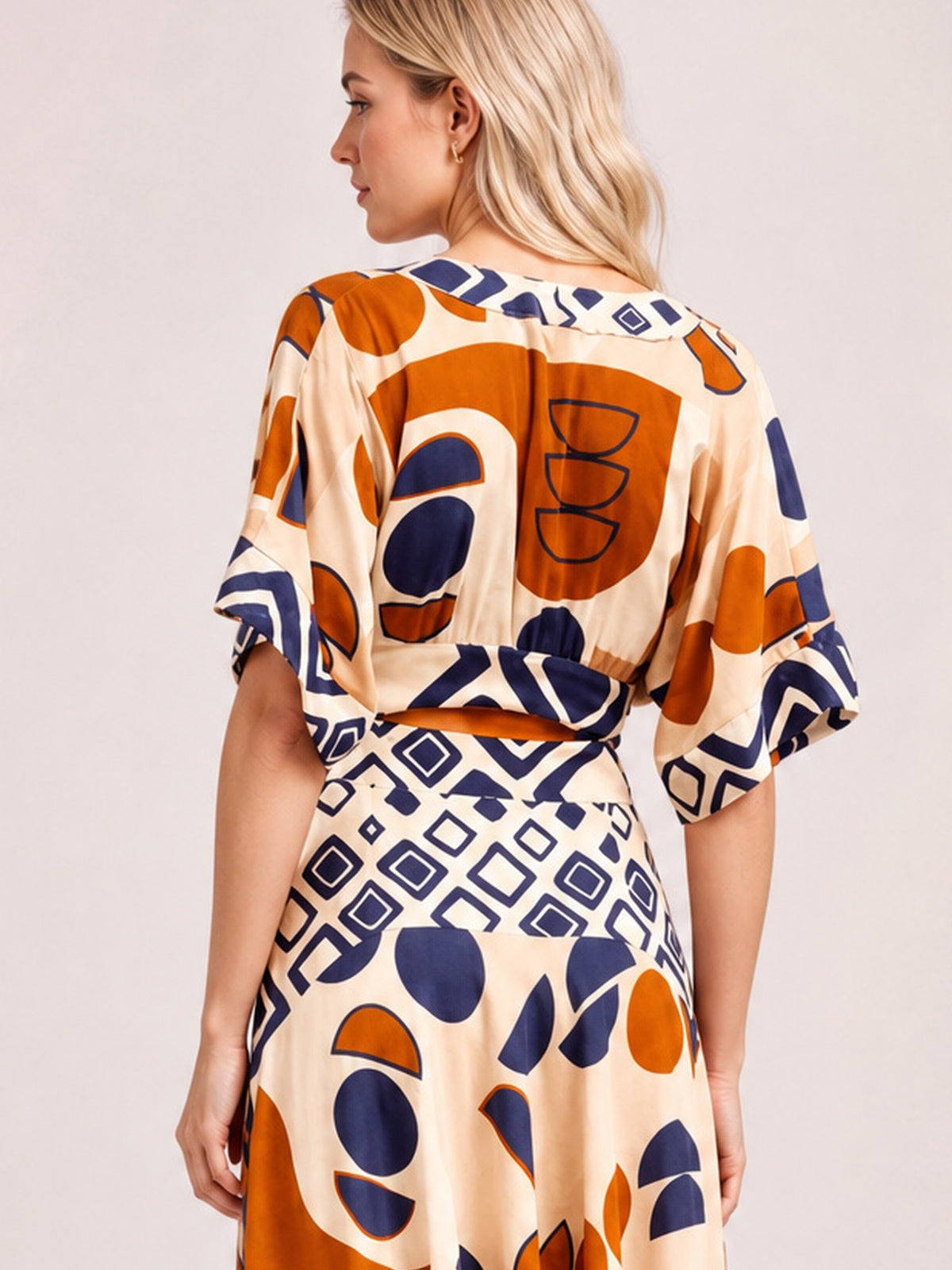 Back view of a model in a caramel geometric print maxi dress with navy and orange motifs, short sleeves, a defined waist, and flowing floor-length silhouette.
